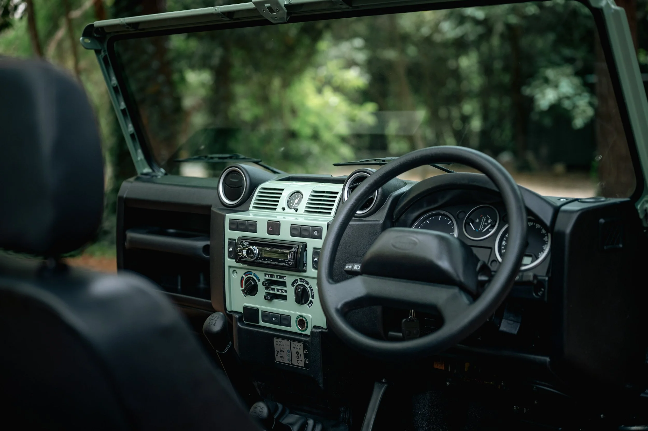 Inside of a vehicle, showing the dashboard and steering wheel, with a view of trees and greenery outside through the windshield.