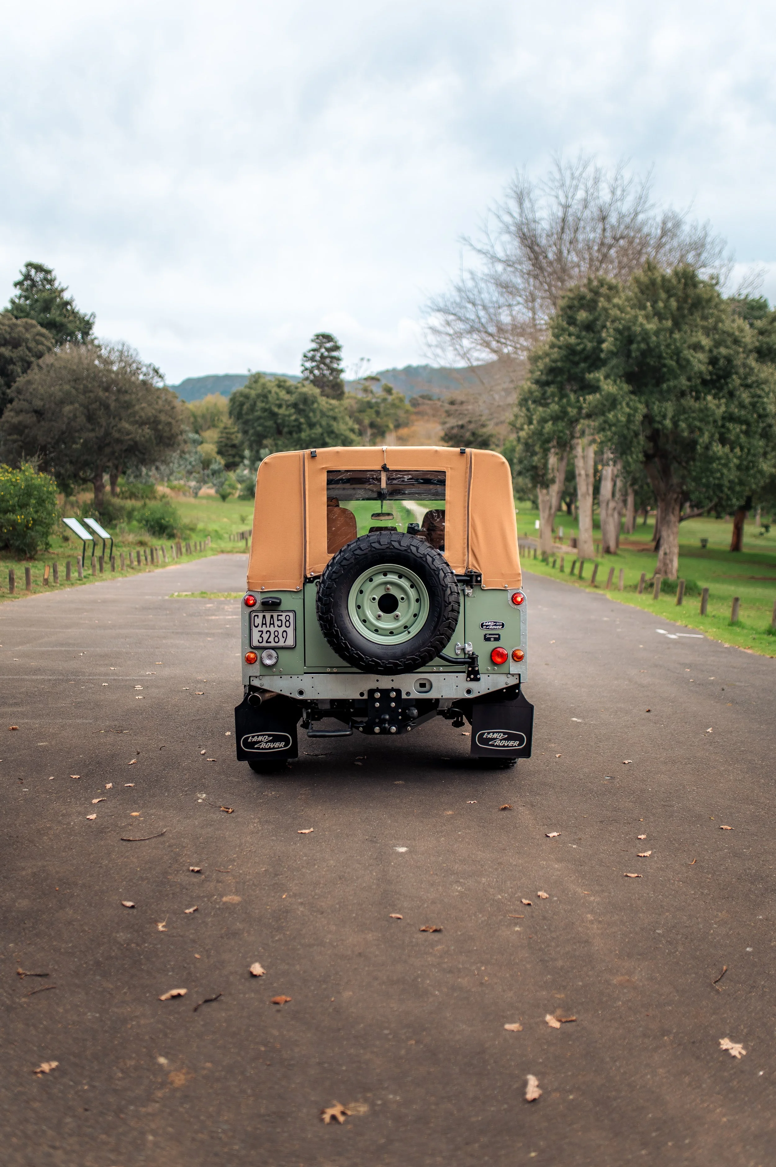Back view of a vintage Land Rover parked on an empty paved path in a park with trees and green hills in the background.