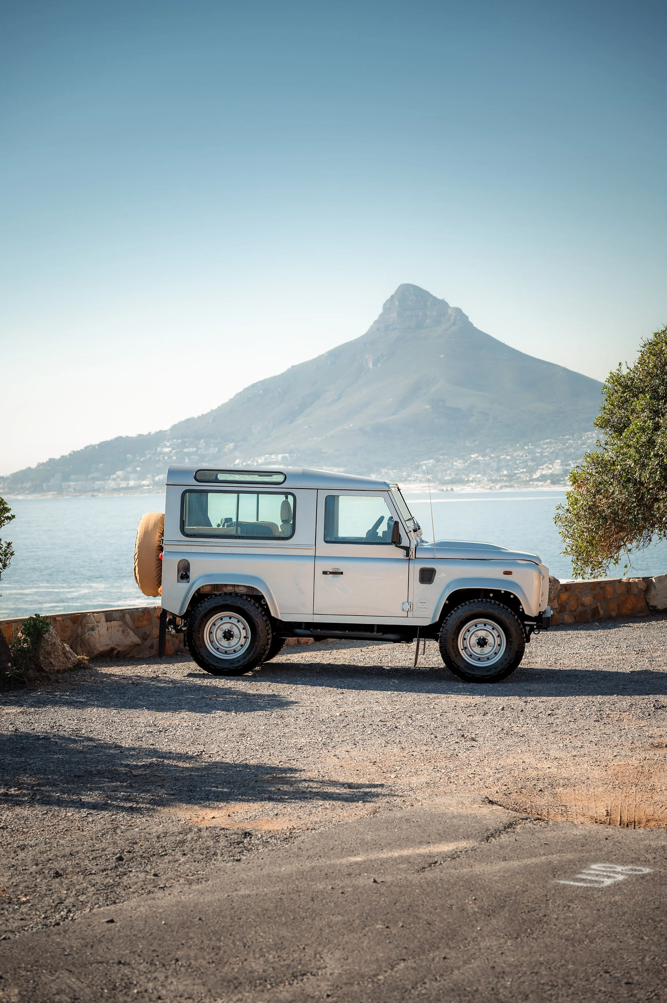 A Zermatt Silver off-road Land rover Defender parked near a body of water in Cape Town, on the Atlantic Seaboard, with a mountain in the background.