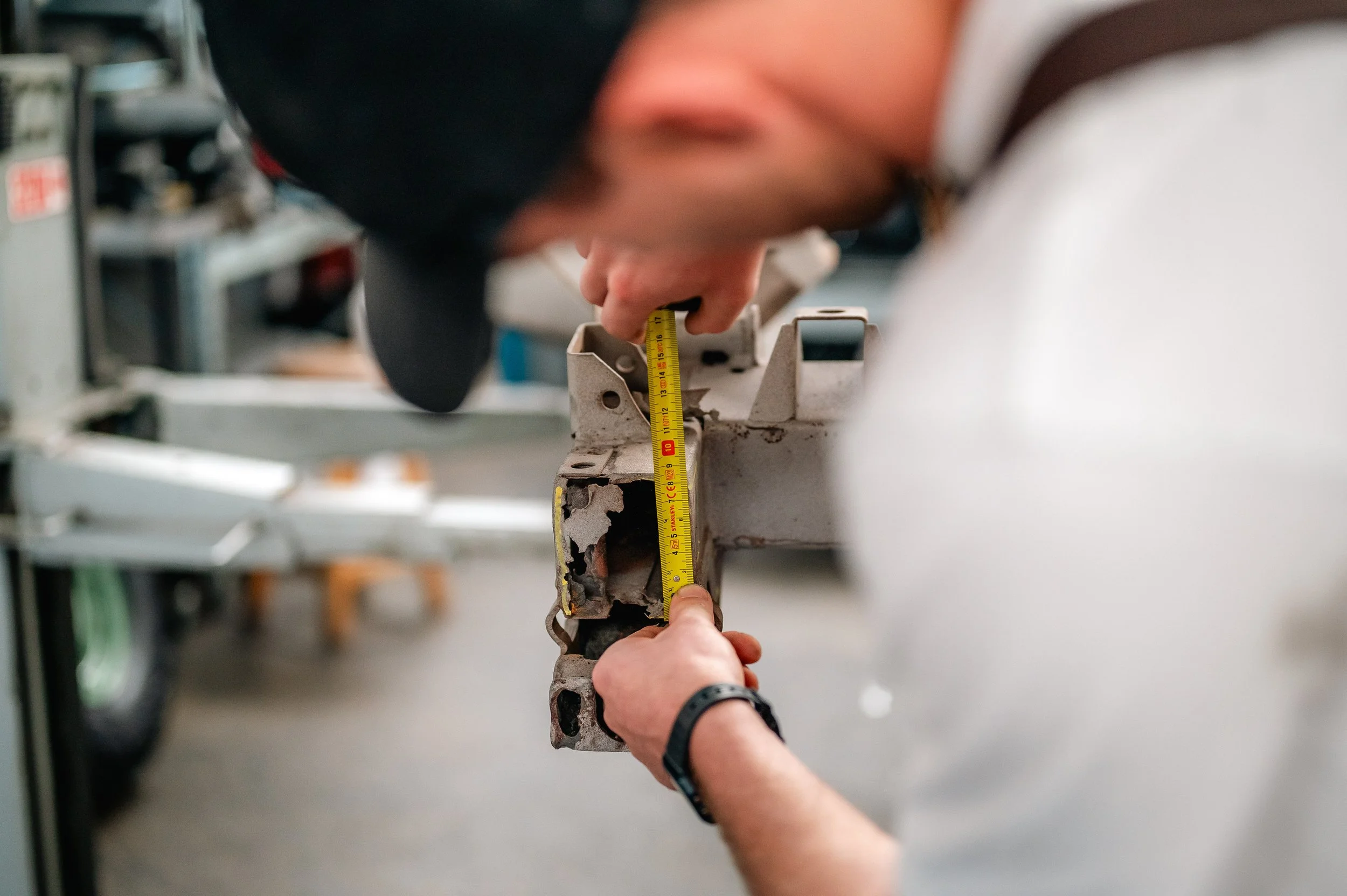 Two men measuring a rusted metal part with a yellow measuring tape in a workshop.
