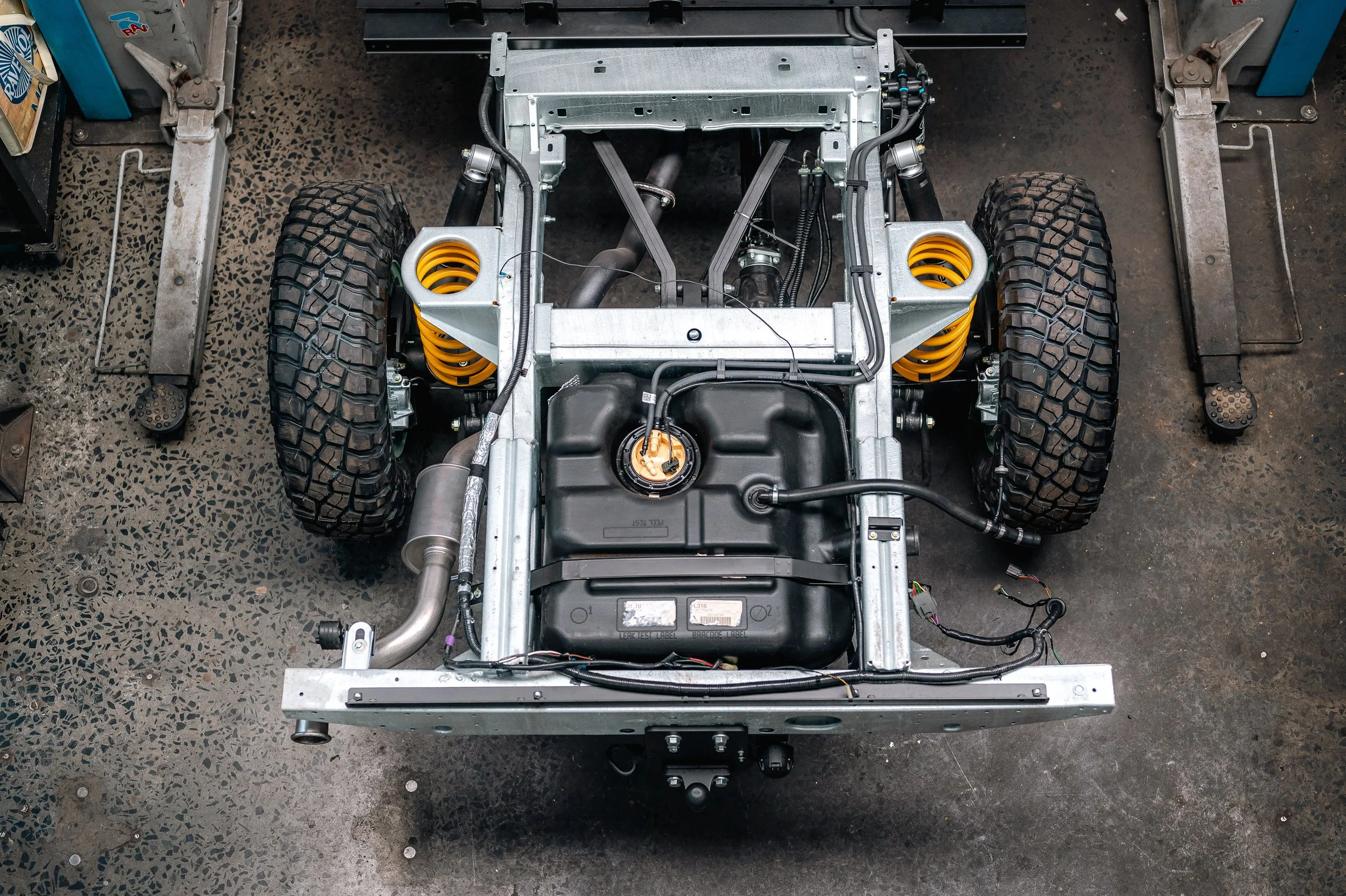 View of a car chassis under construction with large off-road tires, yellow coil springs, and a black fuel tank on a workshop floor.