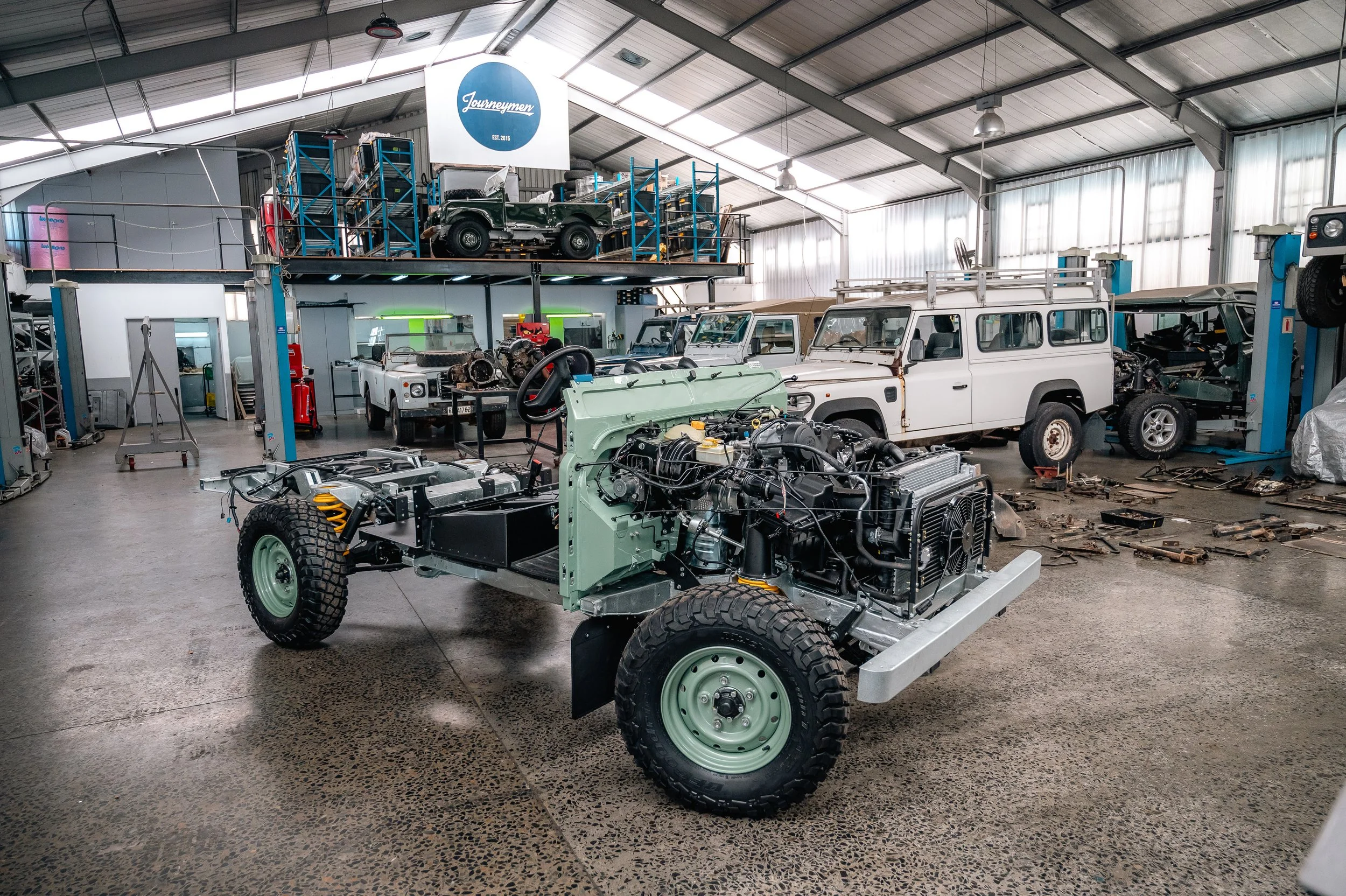 Automotive workshop with vintage and partially assembled vehicles, including a green off-road vehicle in the foreground and a classic white Land Rover in the background.
