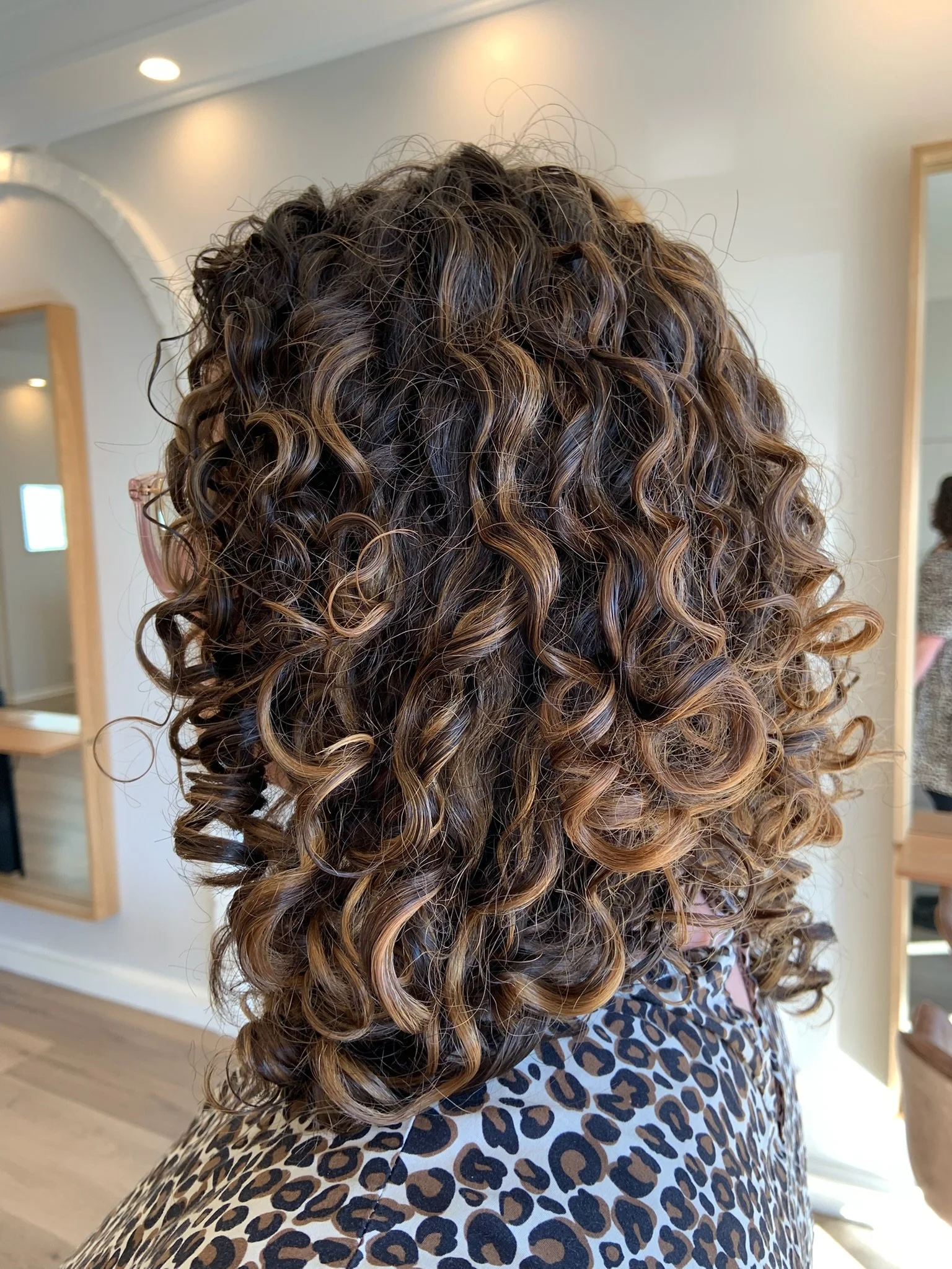 Back view of a person with shoulder-length, curly brown hair with lighter highlights, wearing a floral-patterned top.