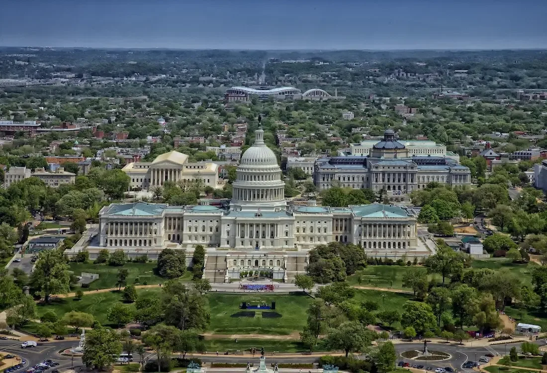 Aerial view of the U.S. Capitol surrounded by green trees and historic buildings under a clear blue sky in Washington, D.C.