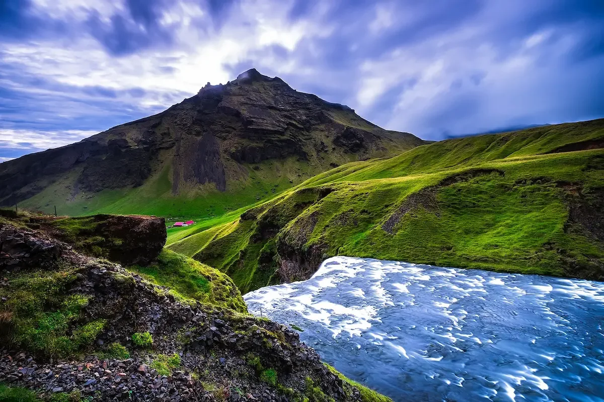 Dramatic mountain landscape with a cascading river in the foreground, vibrant green hills, a small red house, and moody clouds above. Serene and majestic.