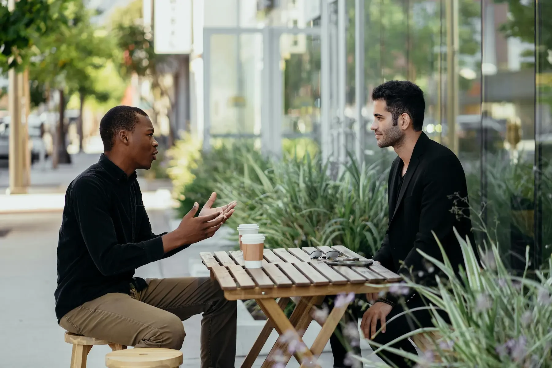 Two men engaged in conversation at a wooden table outside, with coffee cups and greenery surrounding them