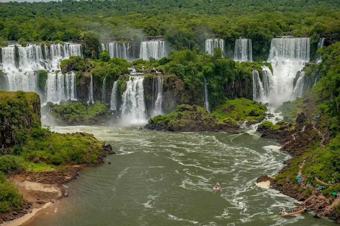 Iguazu Falls cascading over lush green cliffs into a river, with small boats navigating the waters below.