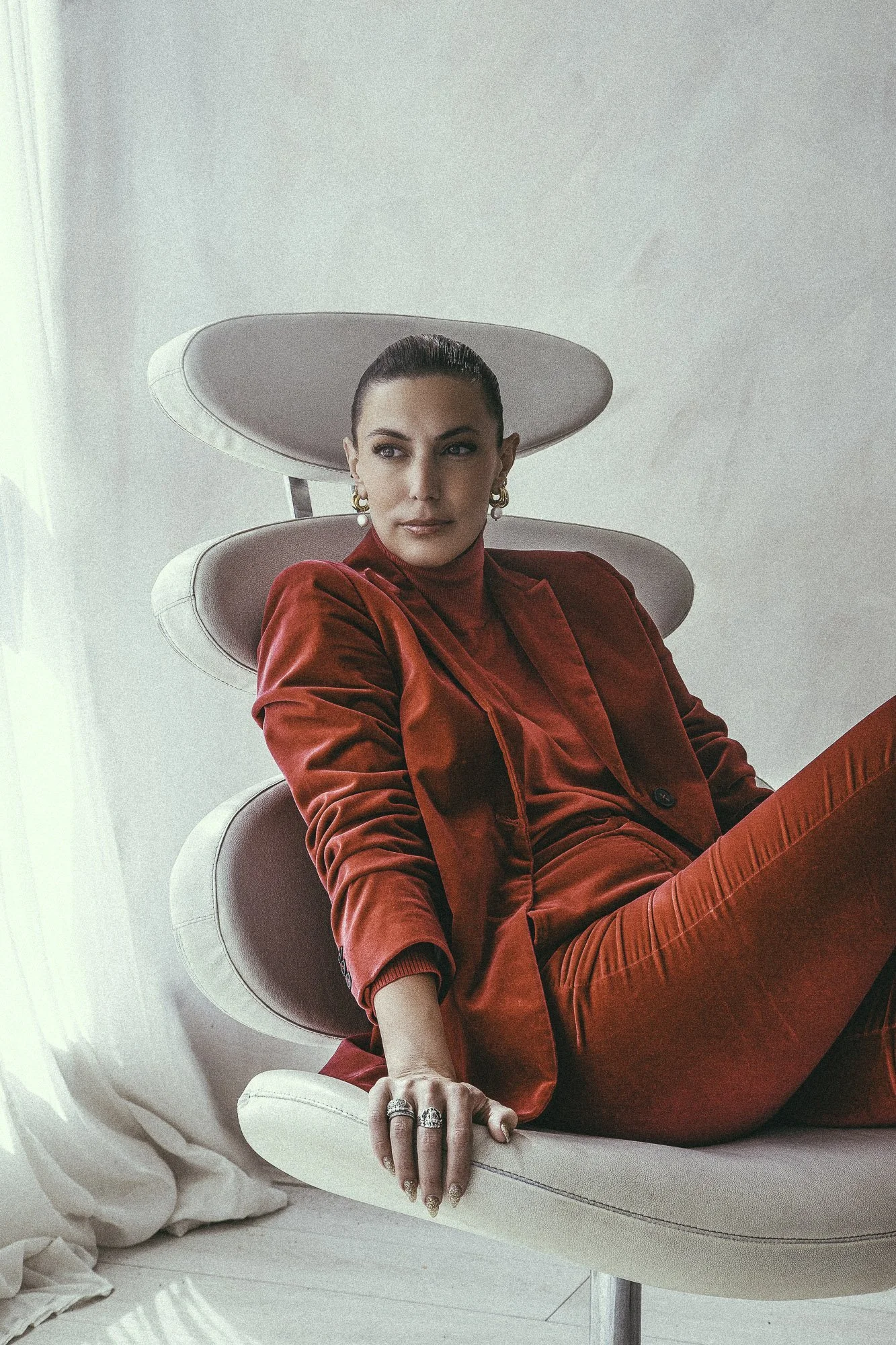 Woman in a red velvet suit sitting on a modern white lounge chair with a unique, layered backrest design, in a well-lit room with white curtains and floor.