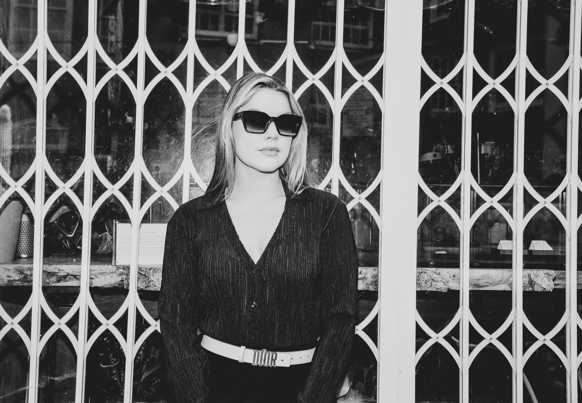 A woman with long hair, wearing dark sunglasses and a dark long-sleeved shirt, standing in front of a metal gate with a geometric pattern.