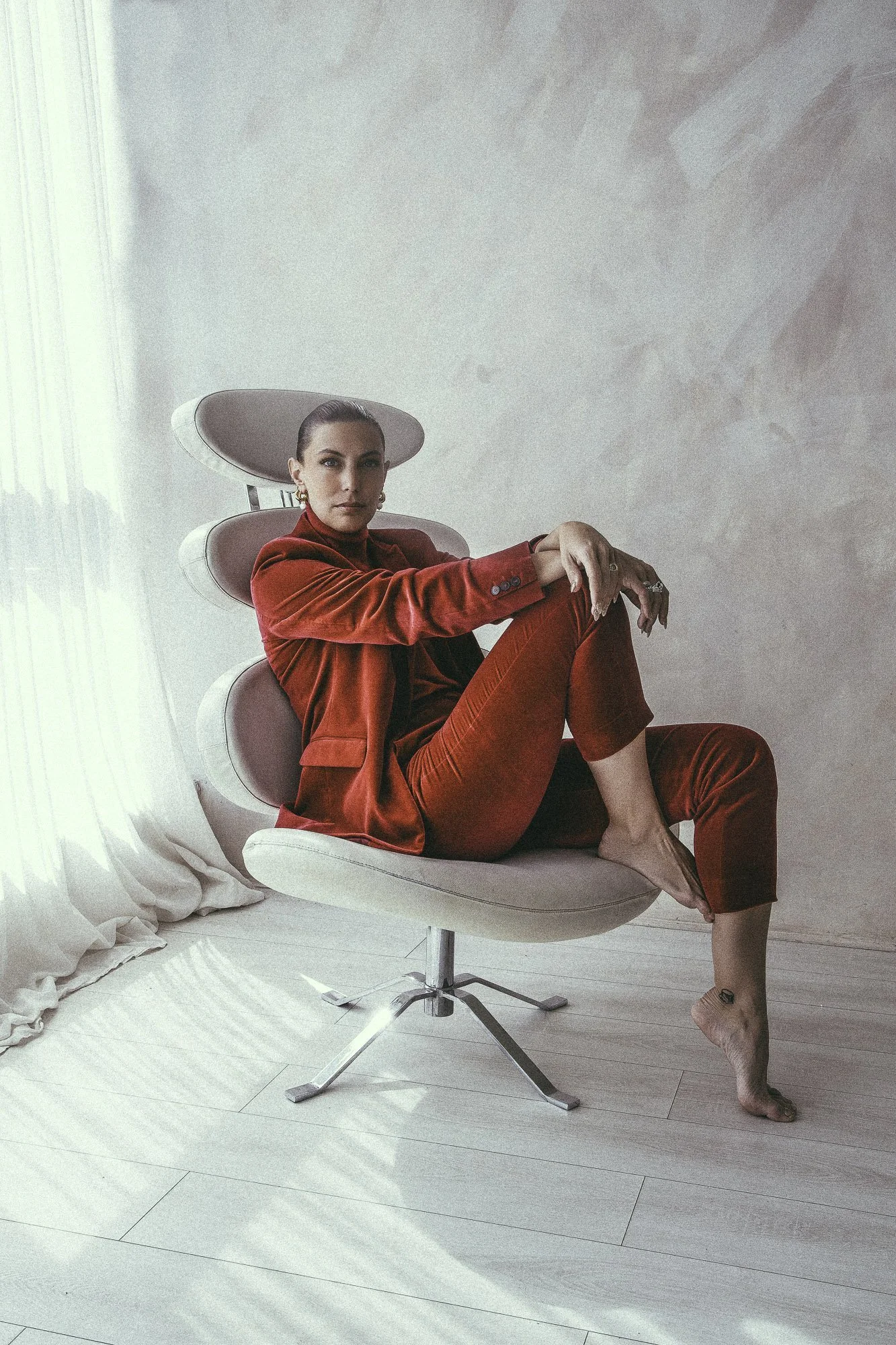 A woman sitting on a pink modern office chair in a minimalist room, dressed in a red suit with a relaxed pose.