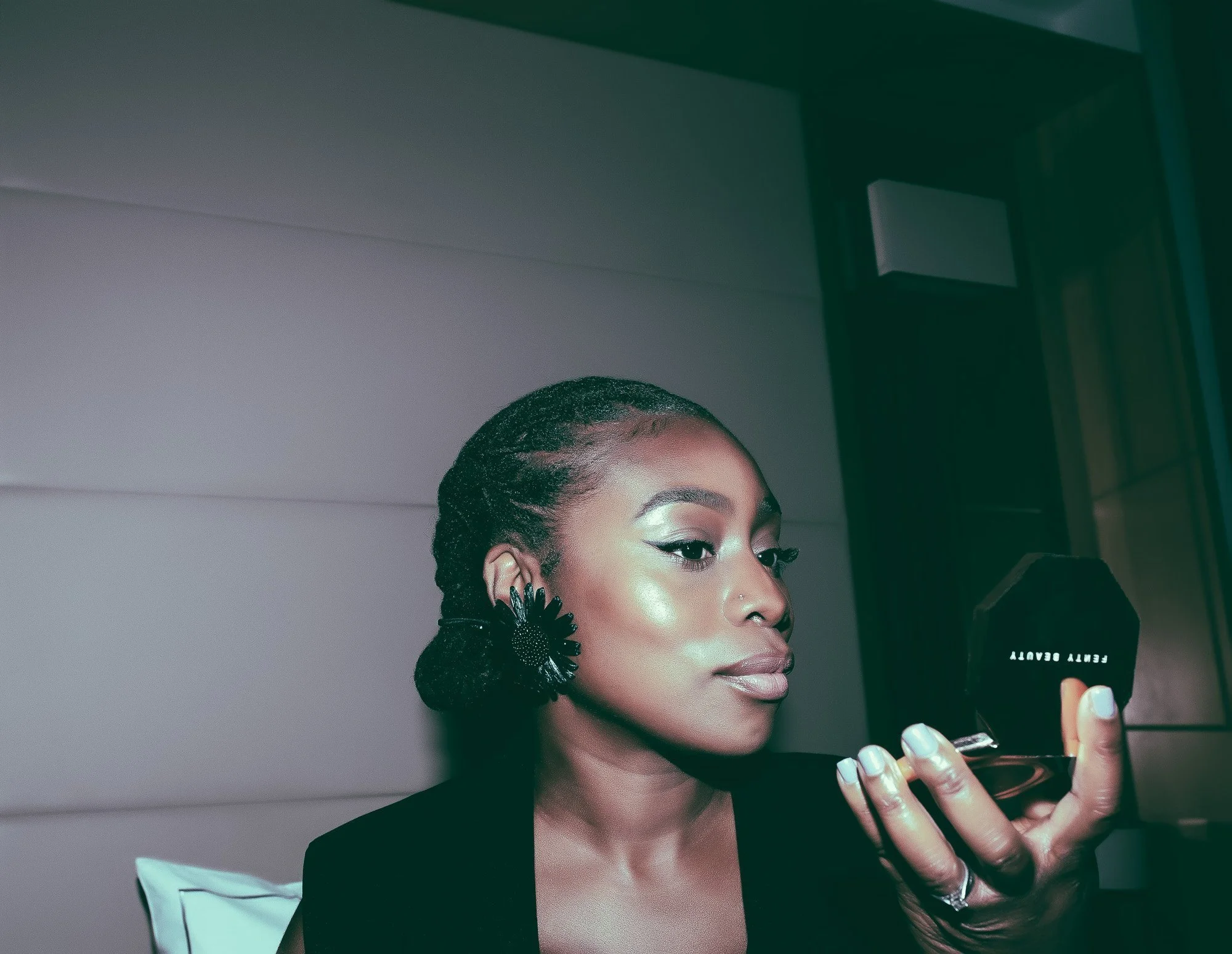 Woman with styled hair and large black earrings looking into a compact mirror labeled 'Beauty Beauty' while applying makeup.
