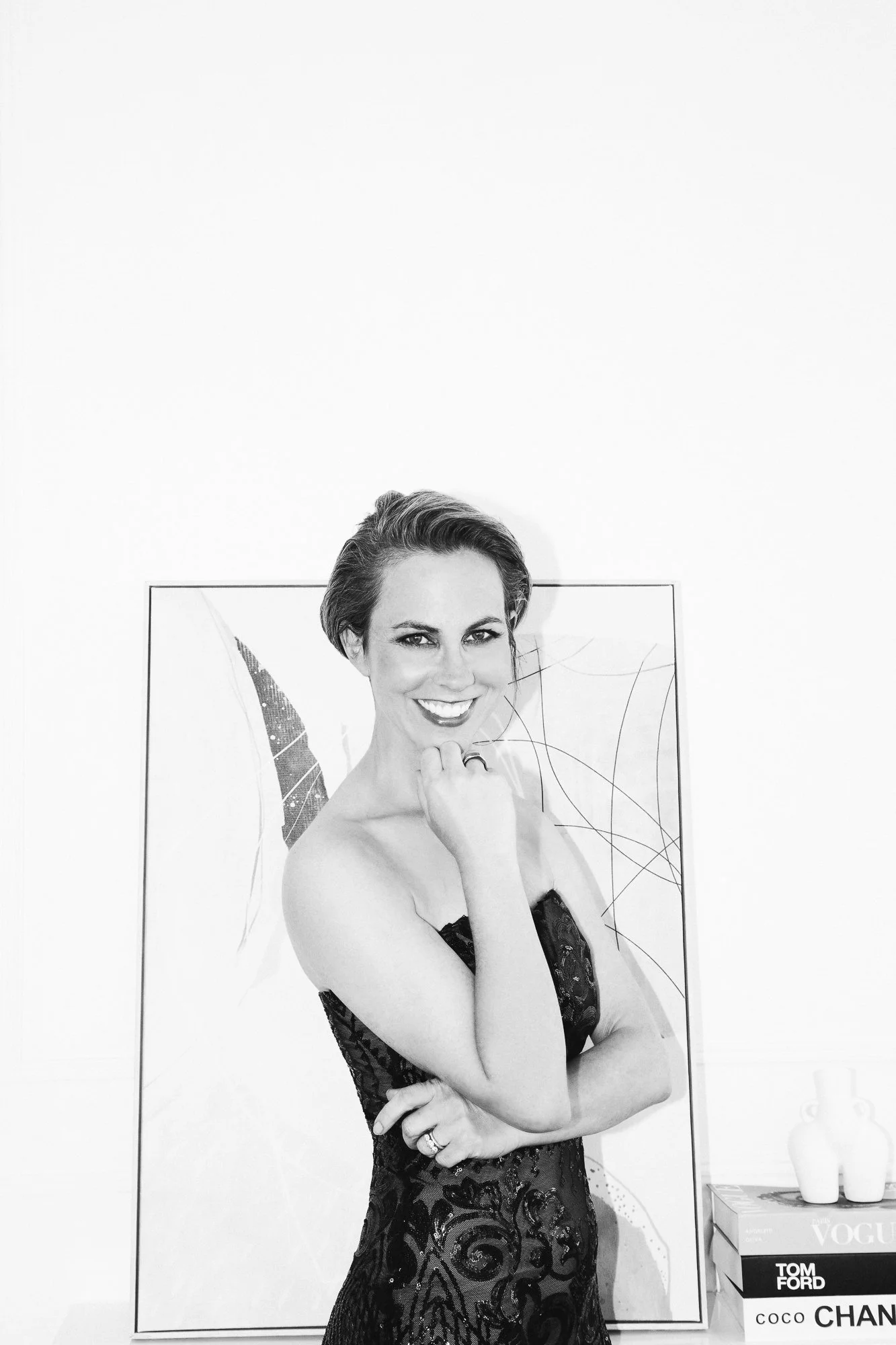 Black and white photo of a smiling woman in a strapless dress, standing with her hand near her chin, in front of an abstract art background.