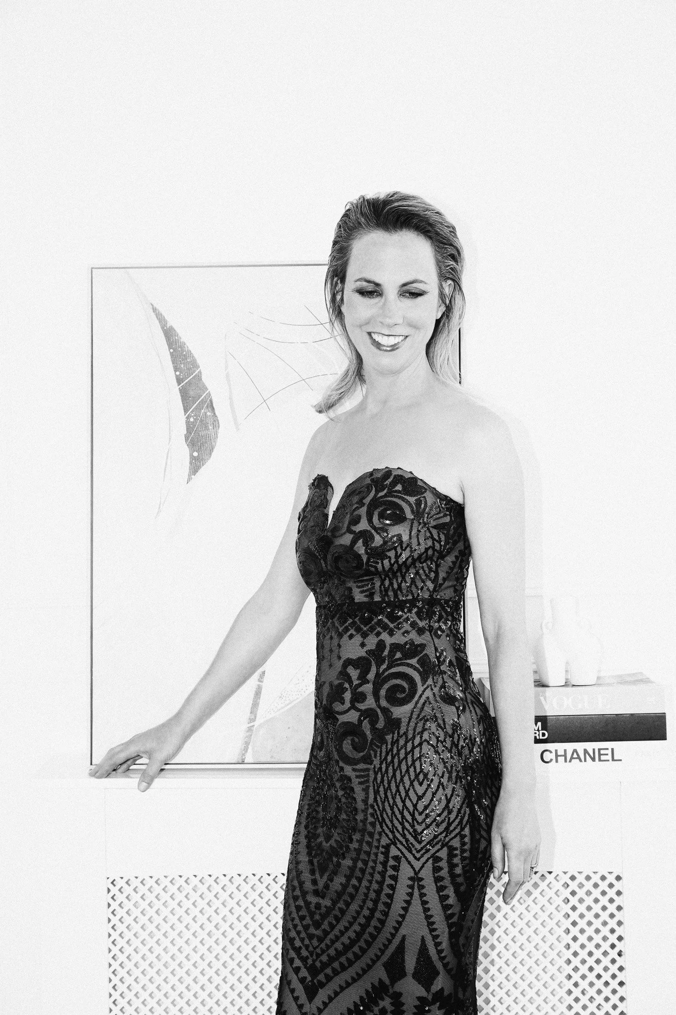 A woman in a strapless black dress standing in front of modern art on a white wall, smiling.