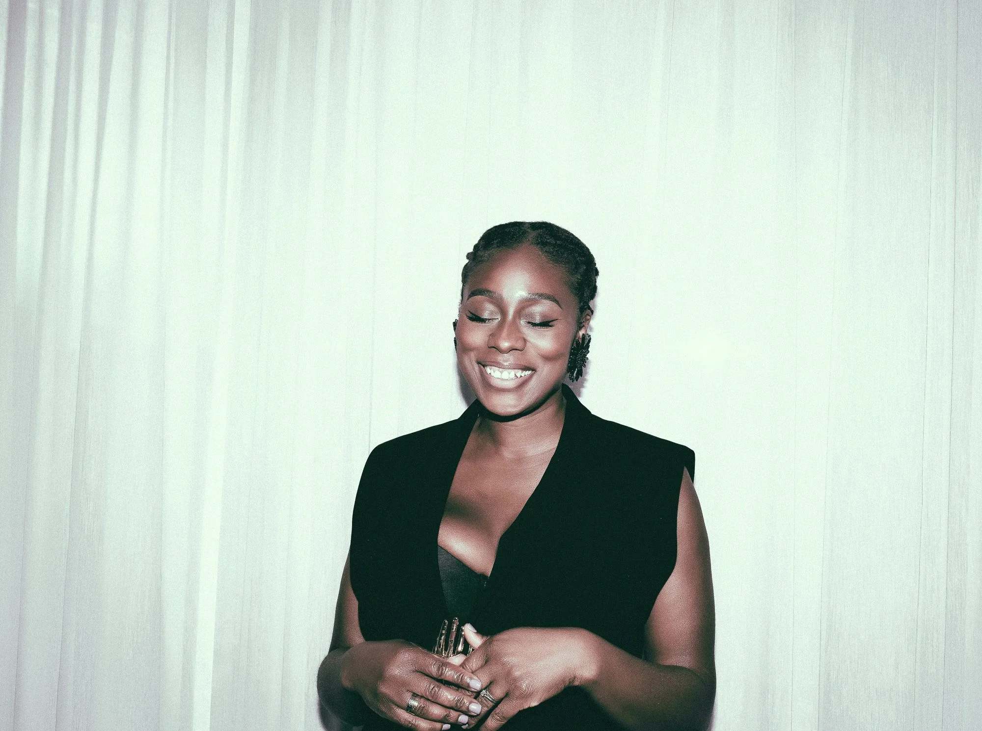 A smiling woman with styled hair and earrings, wearing a sleeveless black outfit, standing against a plain light-colored wall.