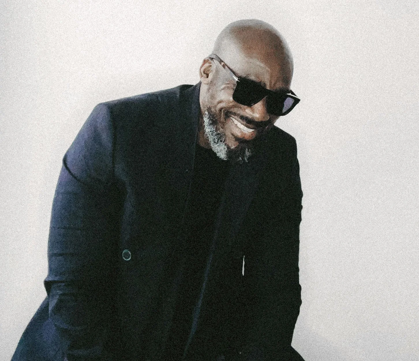 A smiling Black man wearing black sunglasses, a black jacket, and a black shirt, standing against a plain white wall.