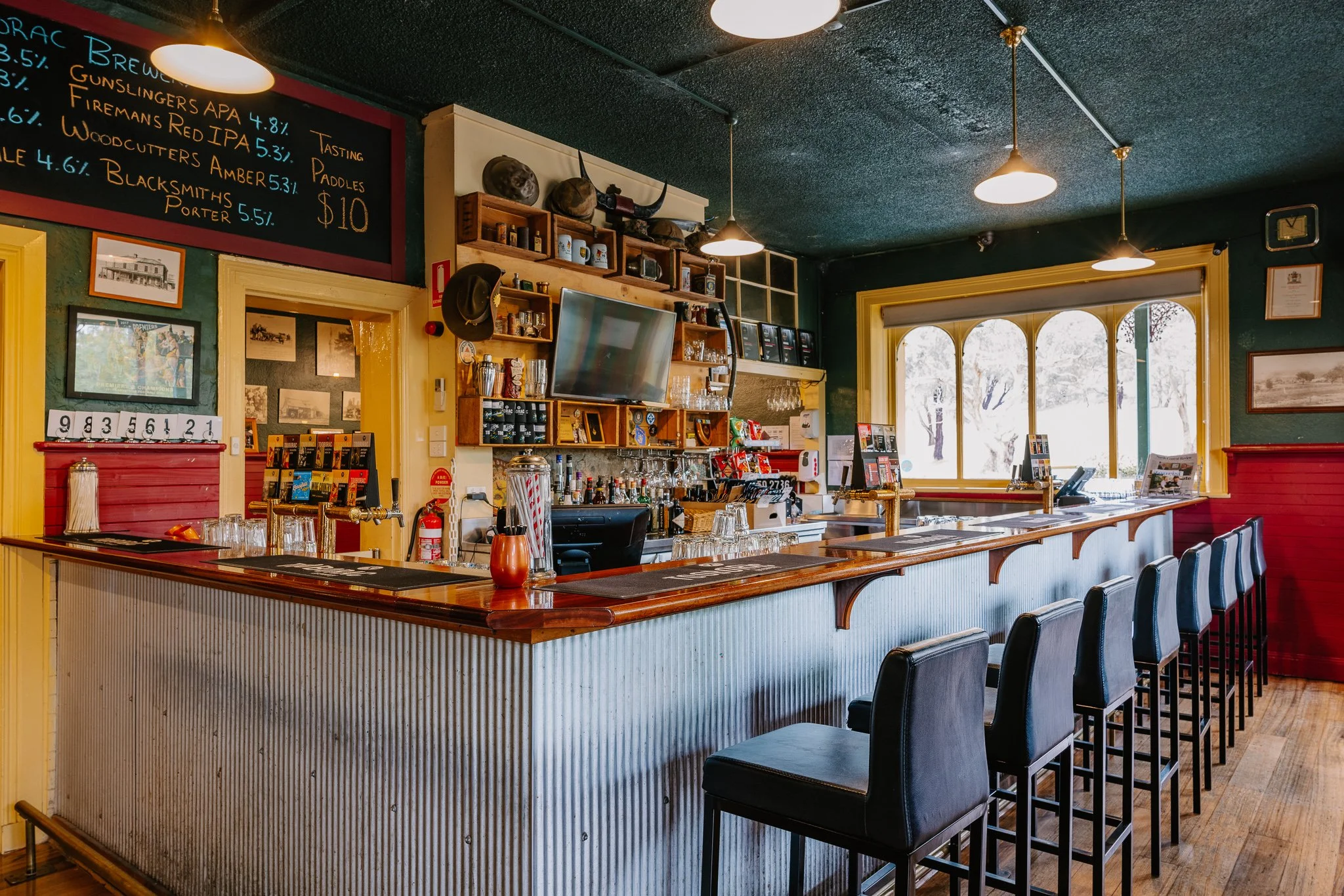 Inside of a cozy bar with a wooden counter, black leather bar stools, a large window letting in natural light, a chalkboard menu displaying beers, shelves with glassware and bottles, and sports memorabilia on the wall.