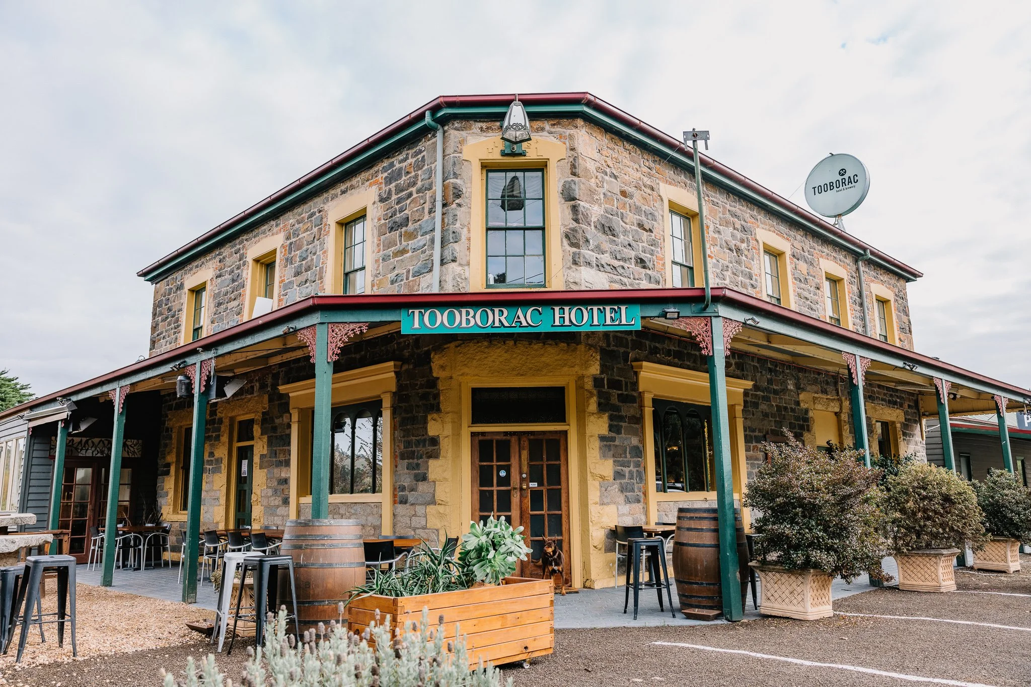 The Tooborac Hotel, a two-story stone building with a green trim and a sign, has outdoor seating with tables, stools, and potted plants in front.