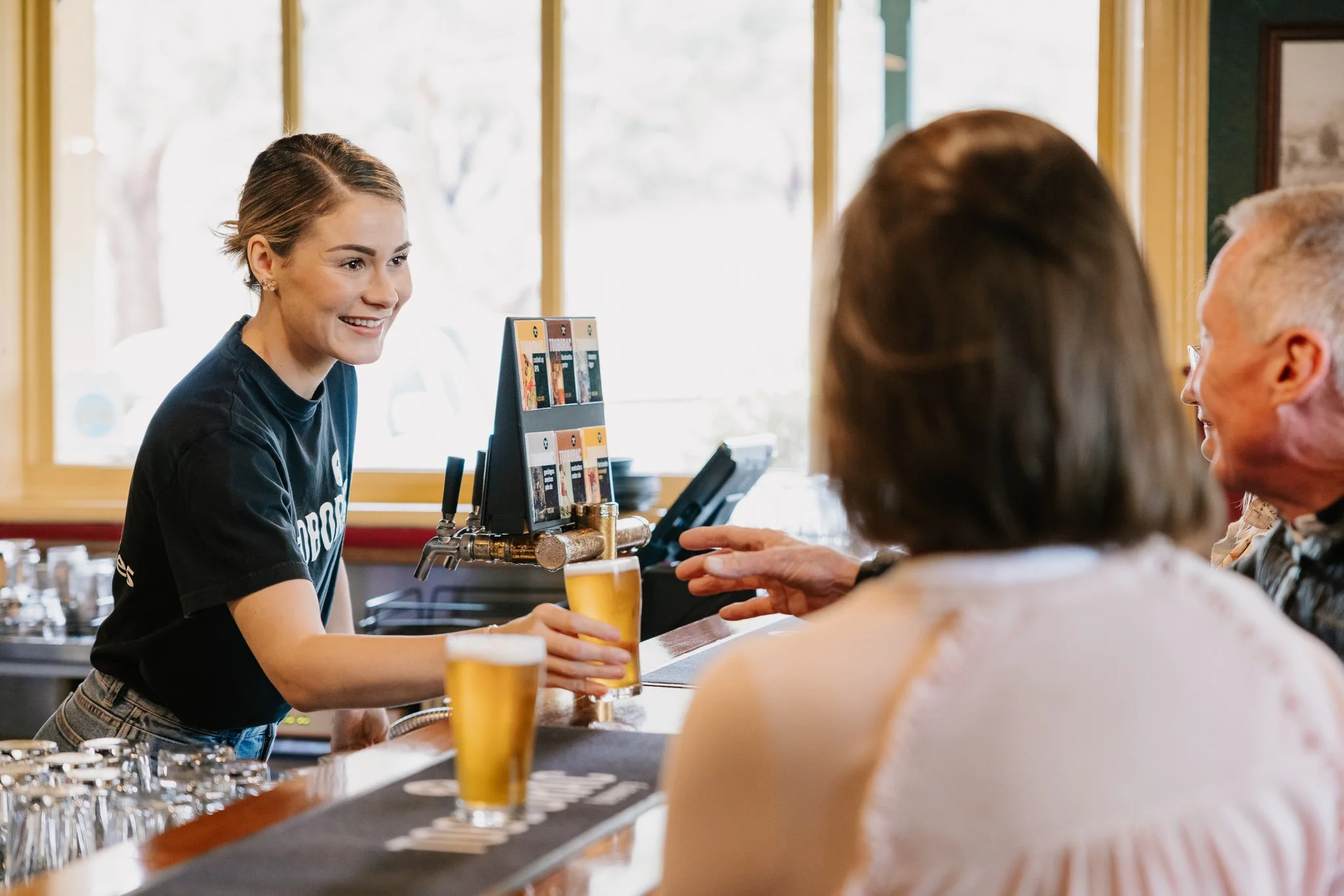 A young woman bartender serving beers to two customers at a bar, with one customer raising a hand for a toast.