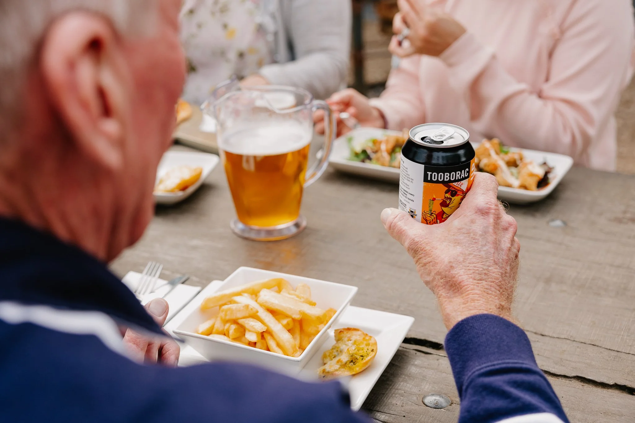 Person holding a can of Toobarac beer at a table with people eating and drinking, including a pitcher of beer and plates of food, outdoors.