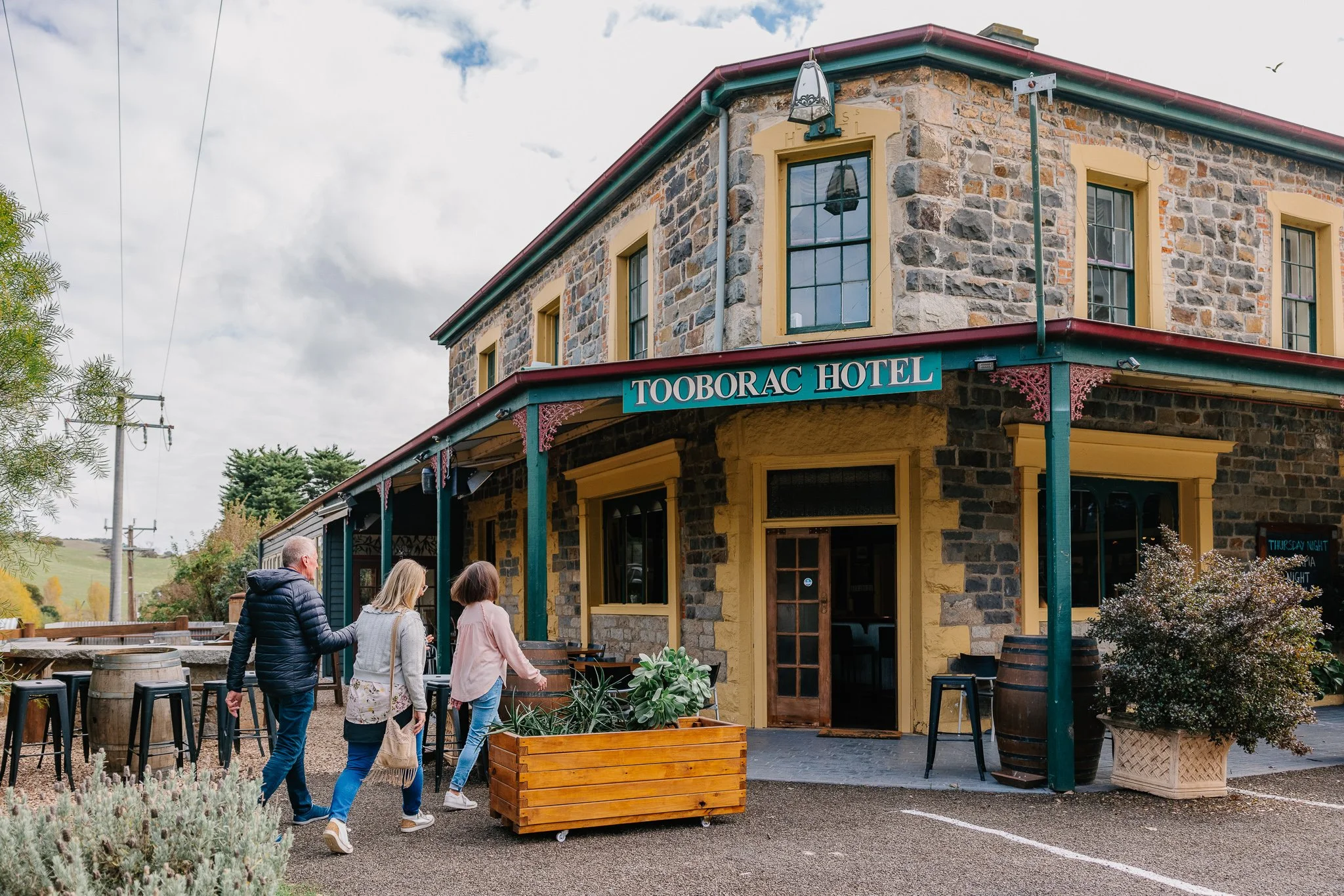 People walking outside Tooborac Hotel, a two-story stone building with a sign, windows with yellow trim, and outdoor seating area.