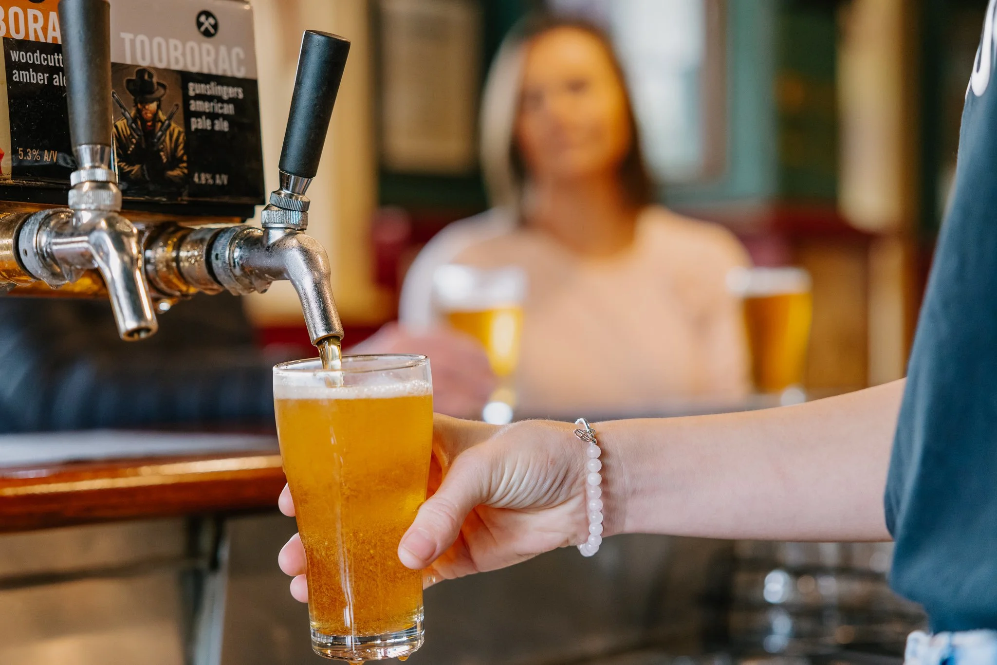 A person pouring a draft beer into a glass at a bar, with other glasses of beer on the bar in the background.