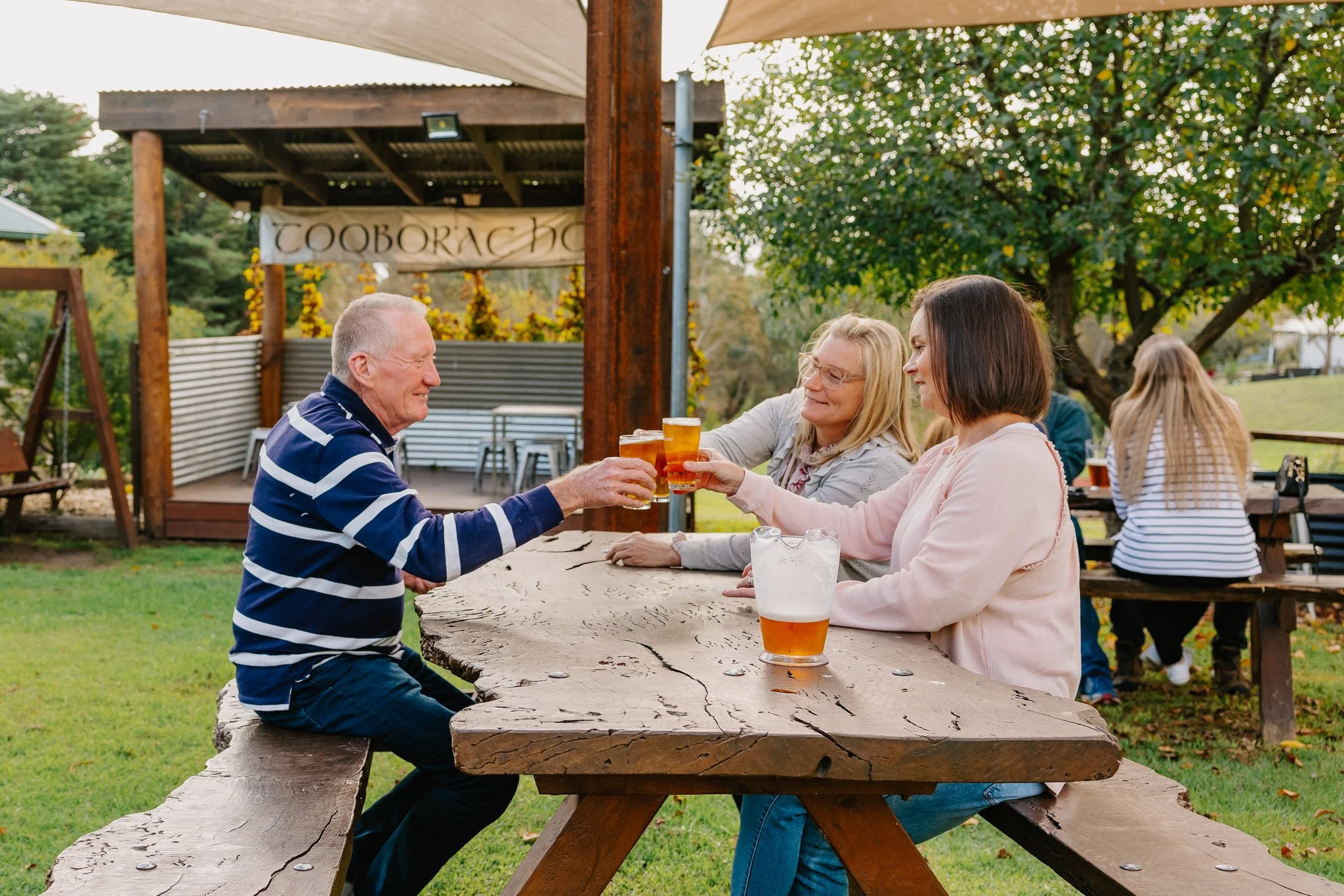 Three people sitting at a wooden picnic table outdoors, clinking glasses of beer, with a woman in a pink jacket, a woman in a light-colored jacket, and an older man in a striped shirt, with other people and trees in the background.