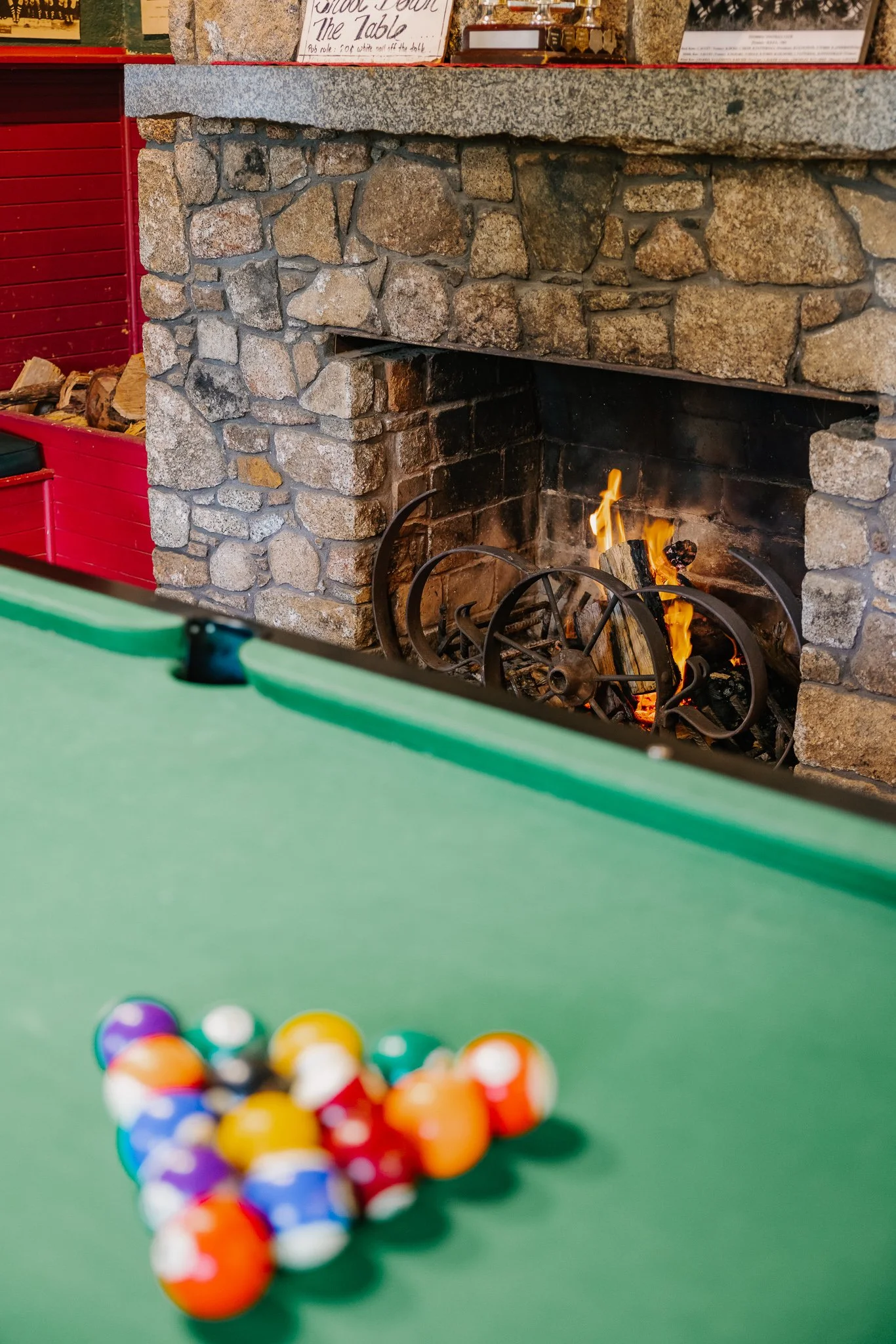 A set of billiard balls arranged in a triangle on a pool table with a stone fireplace and a red wood wall in the background.