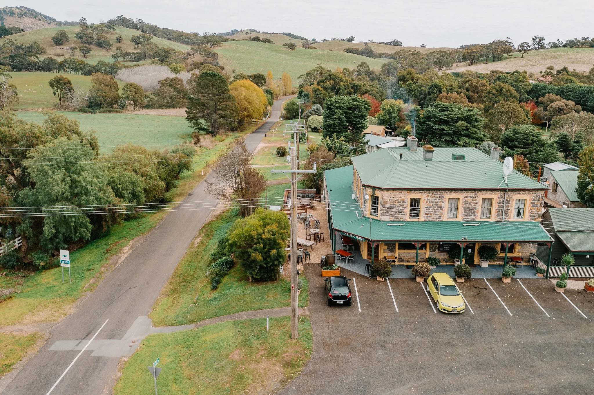 Aerial view of a rural hotel with a green roof, parking lot with cars, outdoor seating, and surrounding green hills and trees.