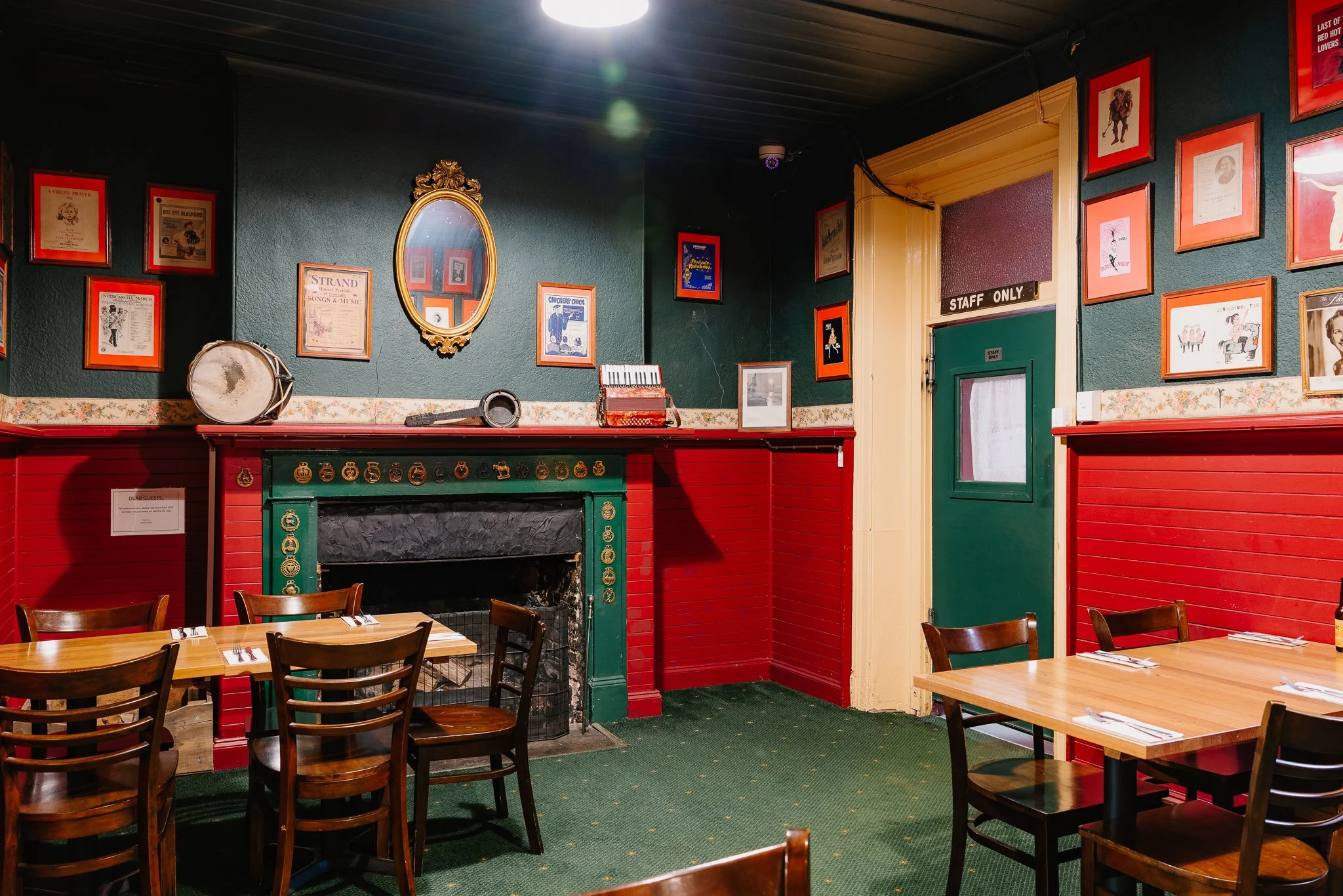 Interior of a cozy restaurant with wooden tables and chairs, green and red walls decorated with framed pictures and memorabilia, and a fireplace with a mirror above it.
