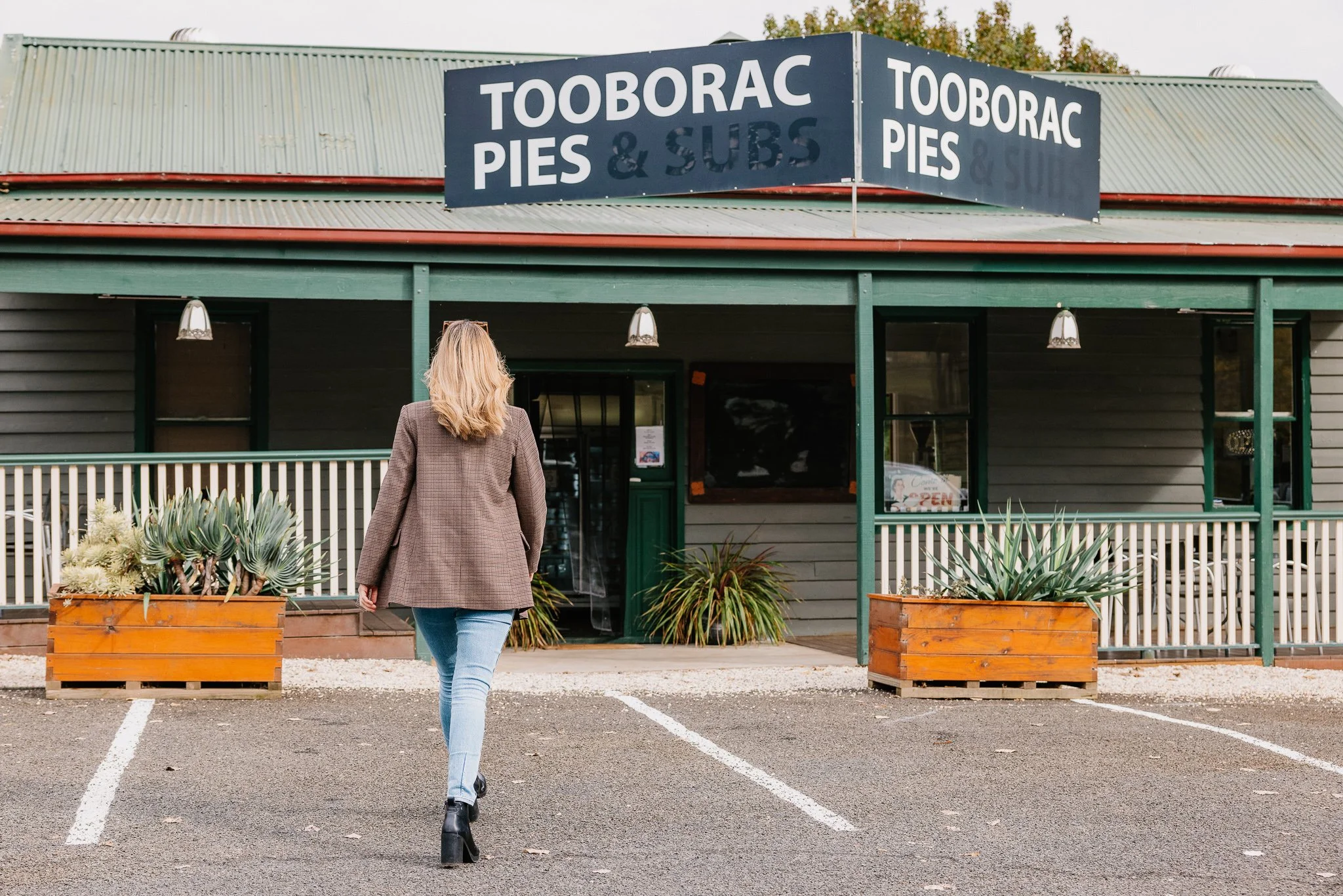 A woman walking toward the entrance of Tooborac Pies & Subs restaurant, with a sign above advertising pies and subs, and outdoor seating with large planter boxes filled with succulents.