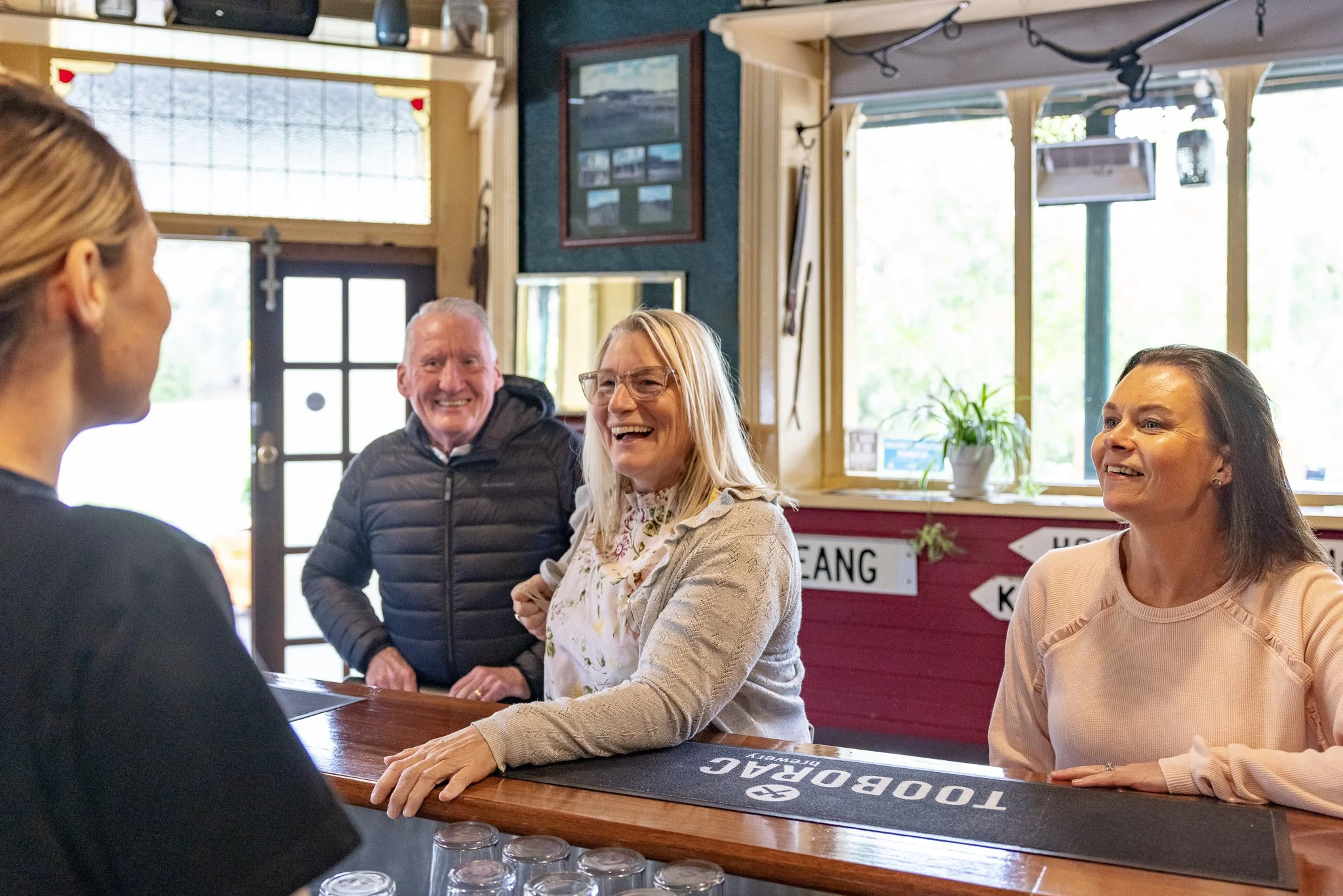 A group of four smiling people at a restaurant bar counter. Two women are seated, and a man stands behind them. The woman on the left is wearing glasses and a light-colored sweater, the woman on the right has dark hair and a light pink top, the man i