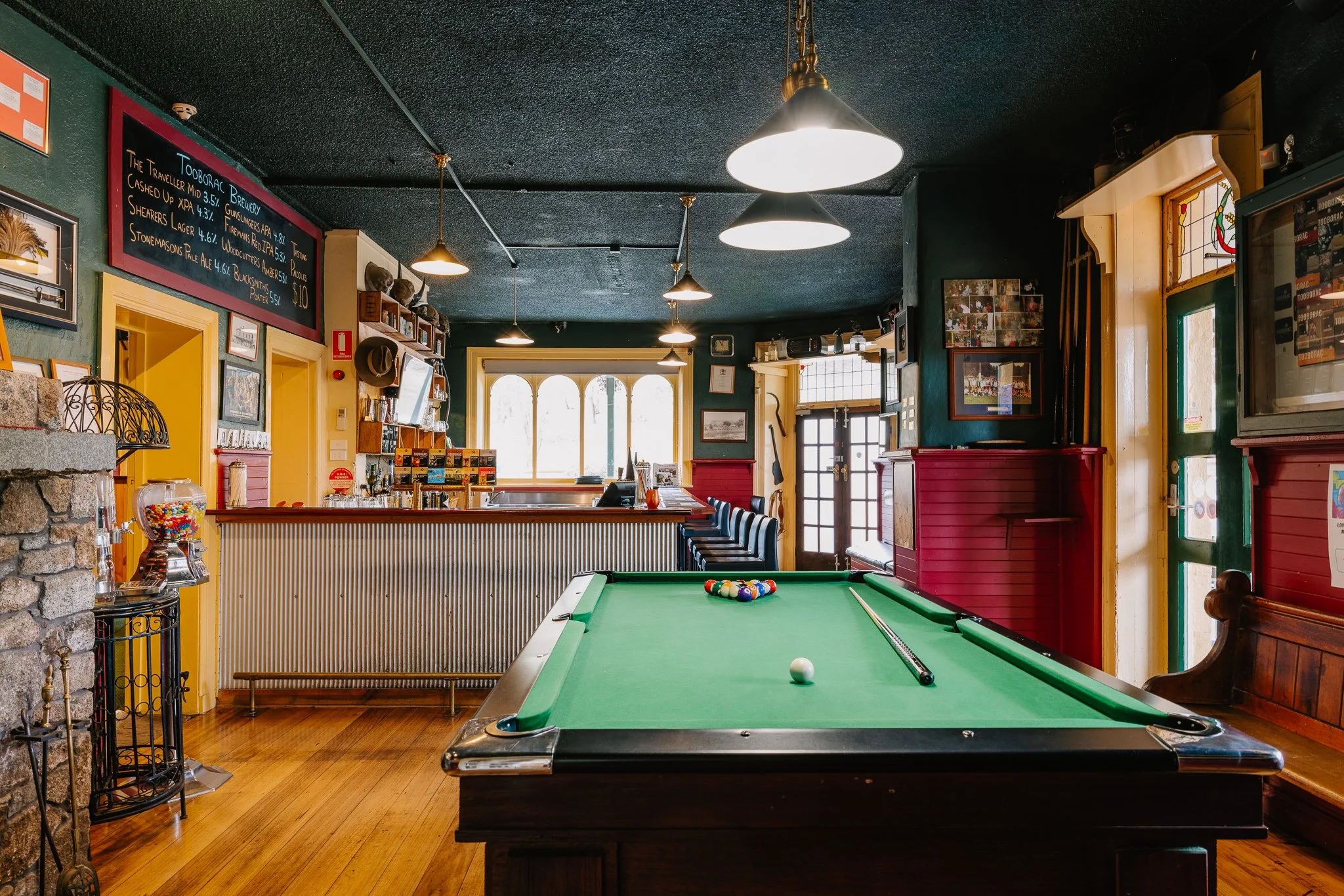 Interior of a cozy bar or pub with a pool table in the foreground, upper lighting fixtures, a bar counter with high chairs, and decorative wall art.