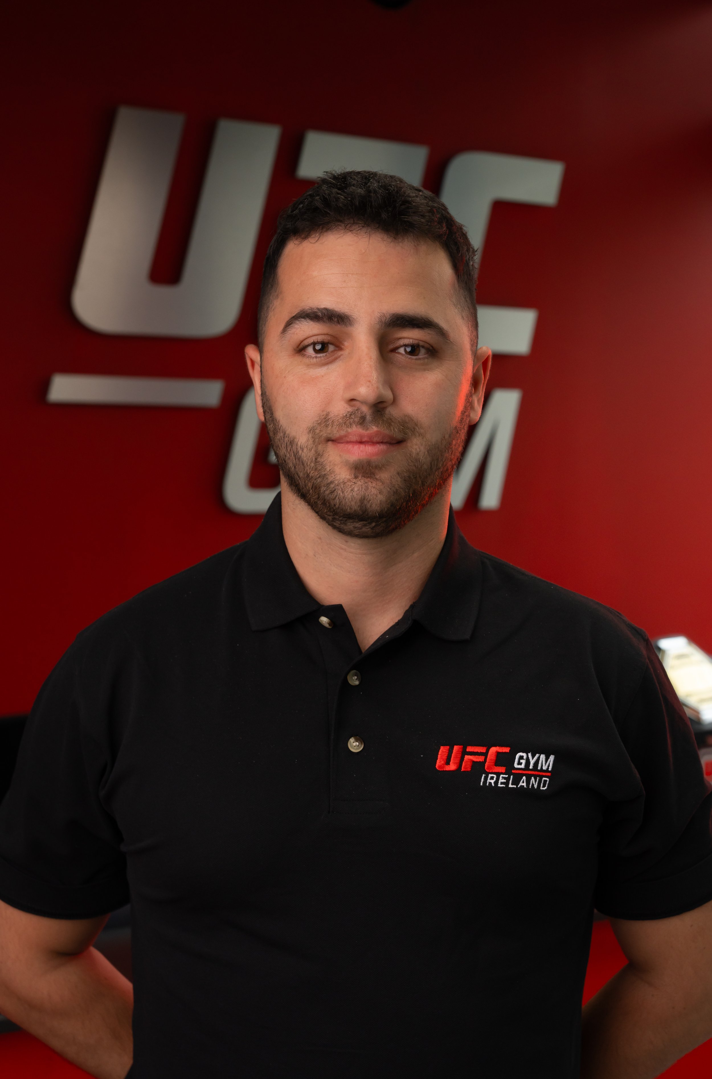 A man wearing a black polo shirt with UFC GYM Ireland logo standing in front of a red wall with large UFC logo.