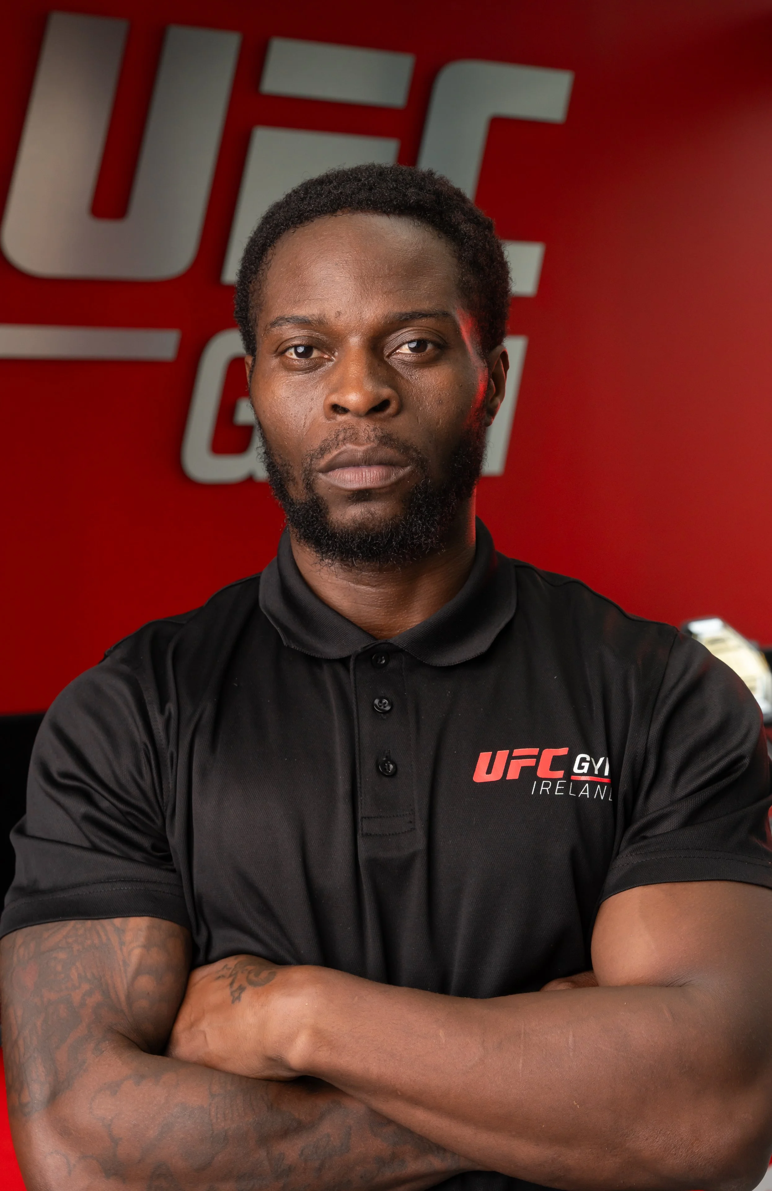 A muscular man with dark curly hair and a beard, wearing a black polo shirt with the UFC Gym Ireland logo, sitting with arms crossed in front of a red wall with a UFC logo.