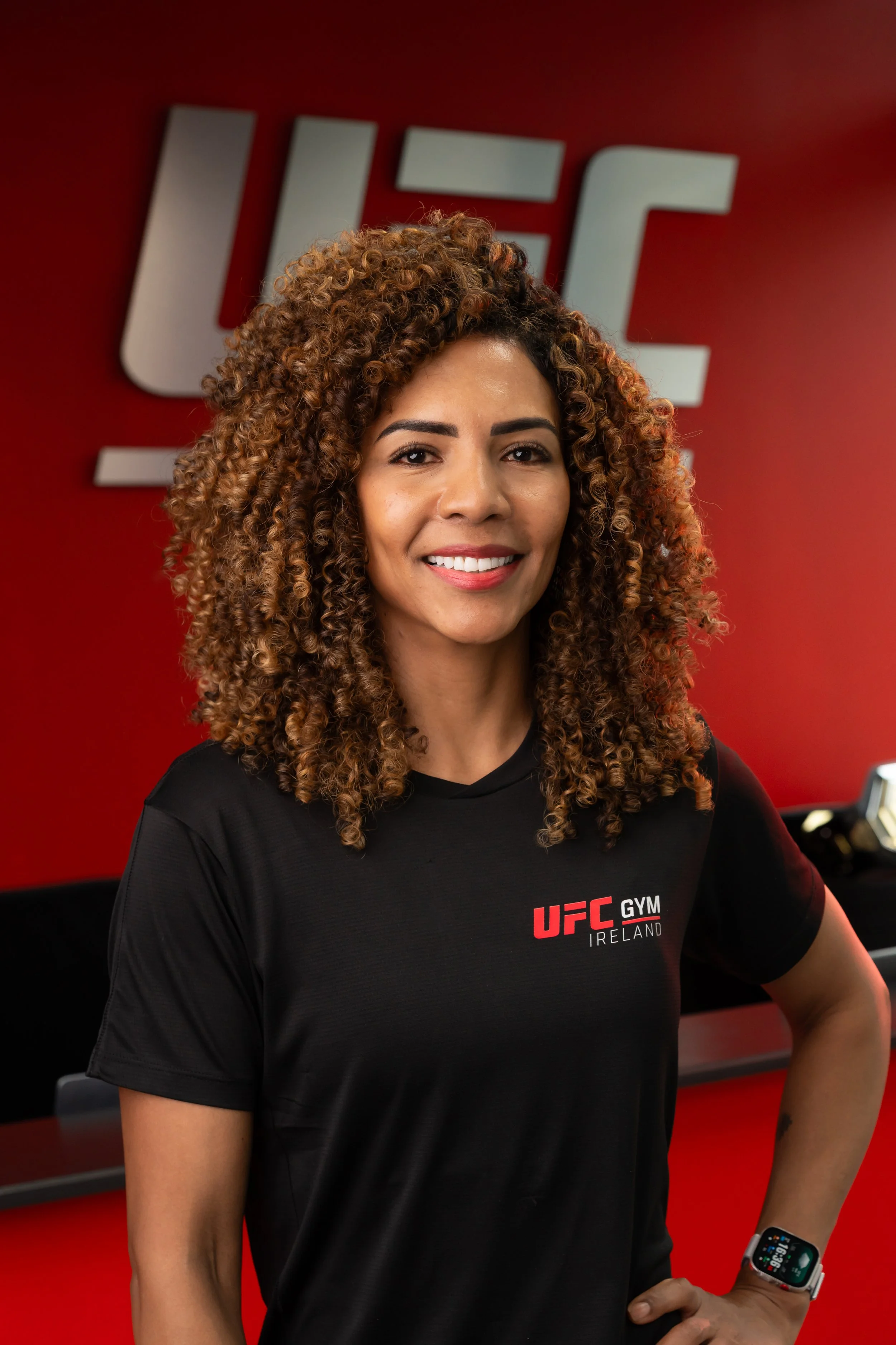 A woman with curly hair smiling, wearing a black UFC Gym Ireland t-shirt and smartwatch, standing in a gym with red walls and UFC logo behind her.