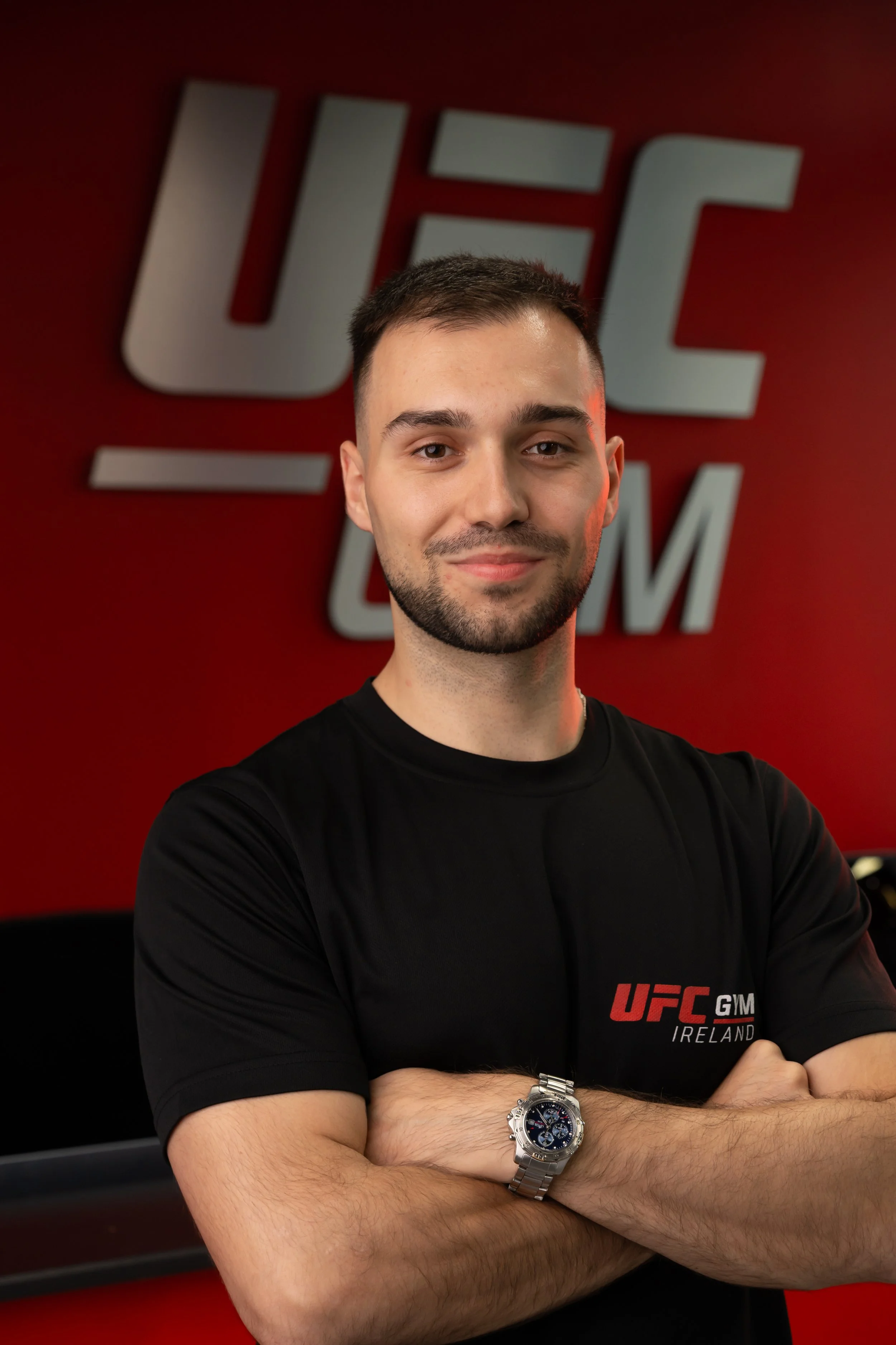 Young man with short dark hair and a beard, wearing a black UFC Ireland shirt and a silver wristwatch, standing with arms crossed in front of a red wall with a UFC logo.