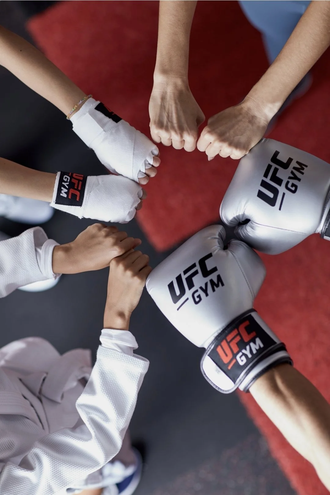 Two people practicing Brazilian Jiu-Jitsu on a red mat, wearing green gis with white belts, face-to-face, with black and white photos hanging on a dark wall behind them.