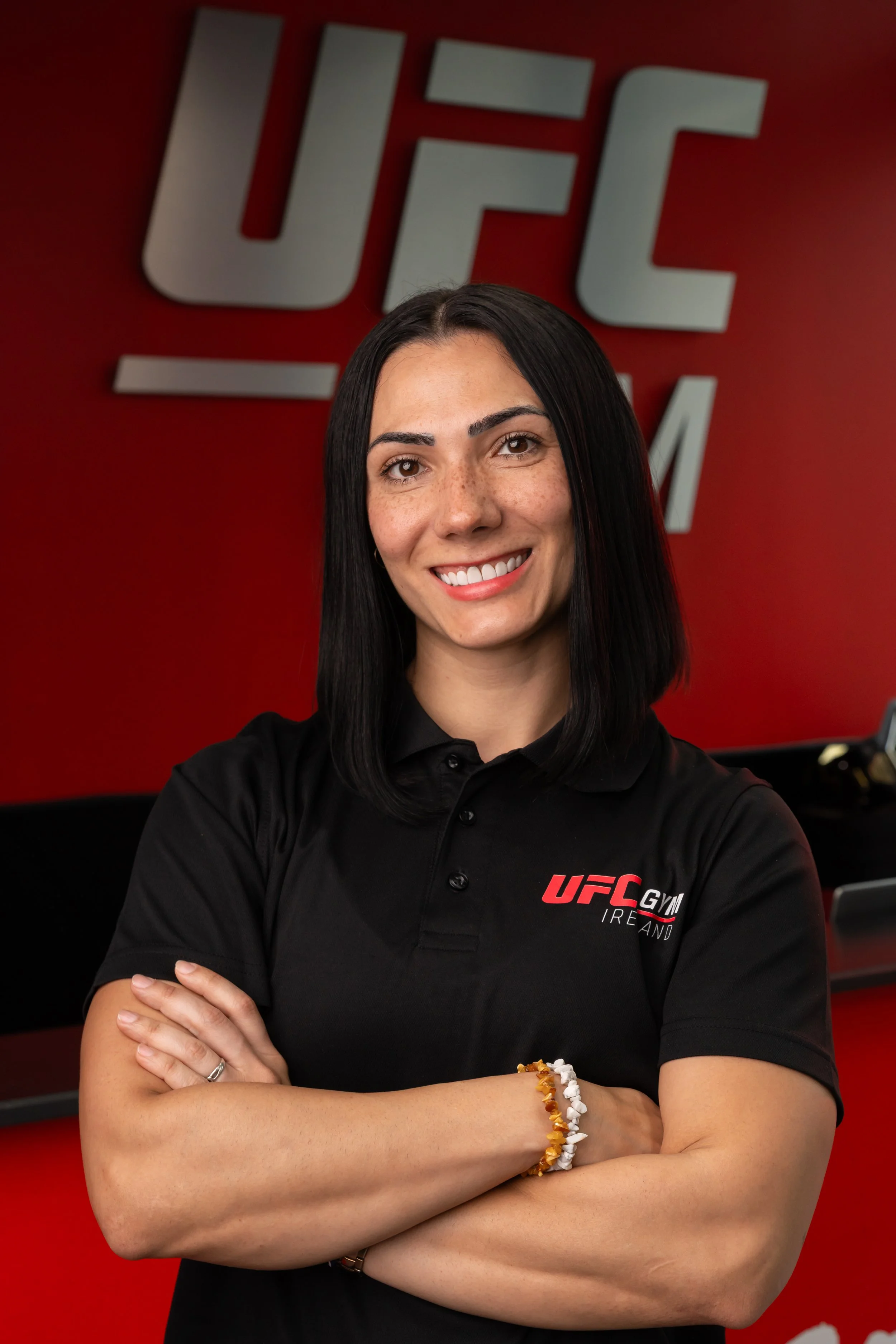 A woman with long black hair, smiling, standing in front of a red wall with a large UFC logo, wearing a black collared shirt with UFC Gym Ireland logo. She has her arms crossed and wears a yellow and white beaded bracelet.