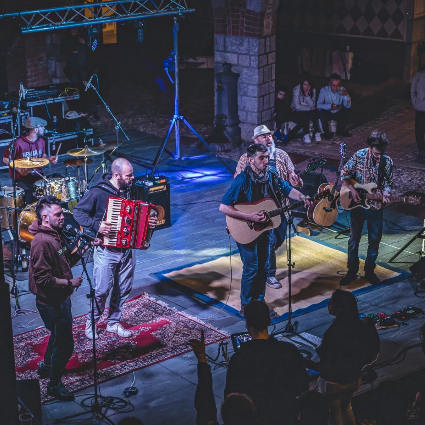 I Menagrama suonano dal vivo su un palco di legno, con strumenti come chitarre, fisarmonica, batteria e microfoni, davanti a un pubblico seduto e alcuni spettatori in piedi, in un ambiente al coperto con pareti di mattoni.