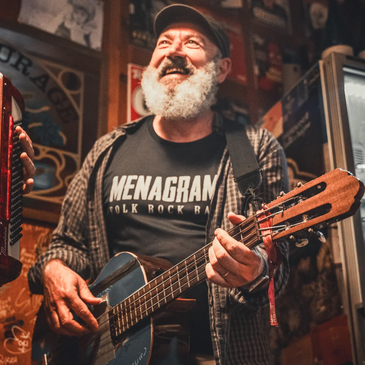 Uomo con barba bianca che suona una chitarra acustica in un bar o locale. Indossa una maglietta nera con scritto 'MENAGRAM' e una camicia a quadri. Sorriso felice, sfondo con decorazioni e scaffali con oggetti.
