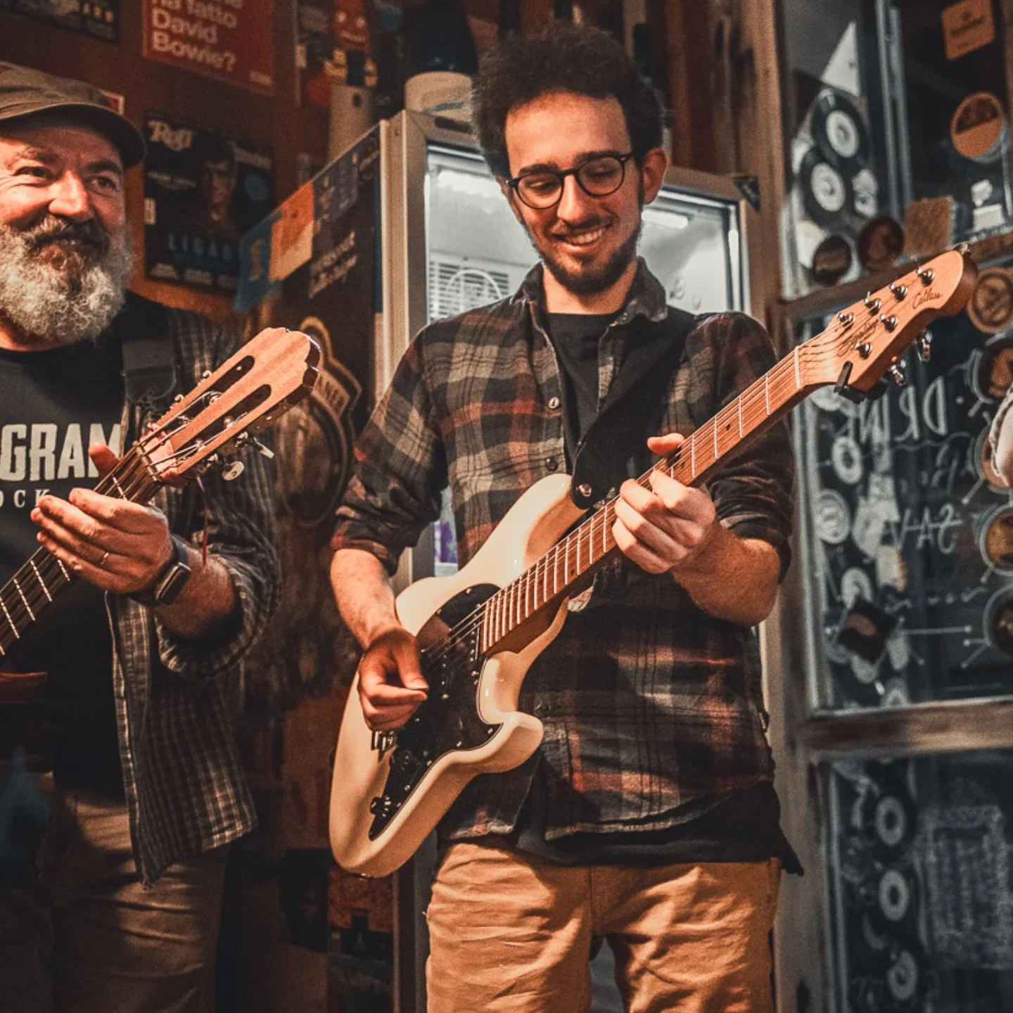 Due uomini suonano chitarre in un locale, uno indossa una camicia a quadri e occhiali, l'altro ha la barba e indossa una maglietta con scritta."Rock".