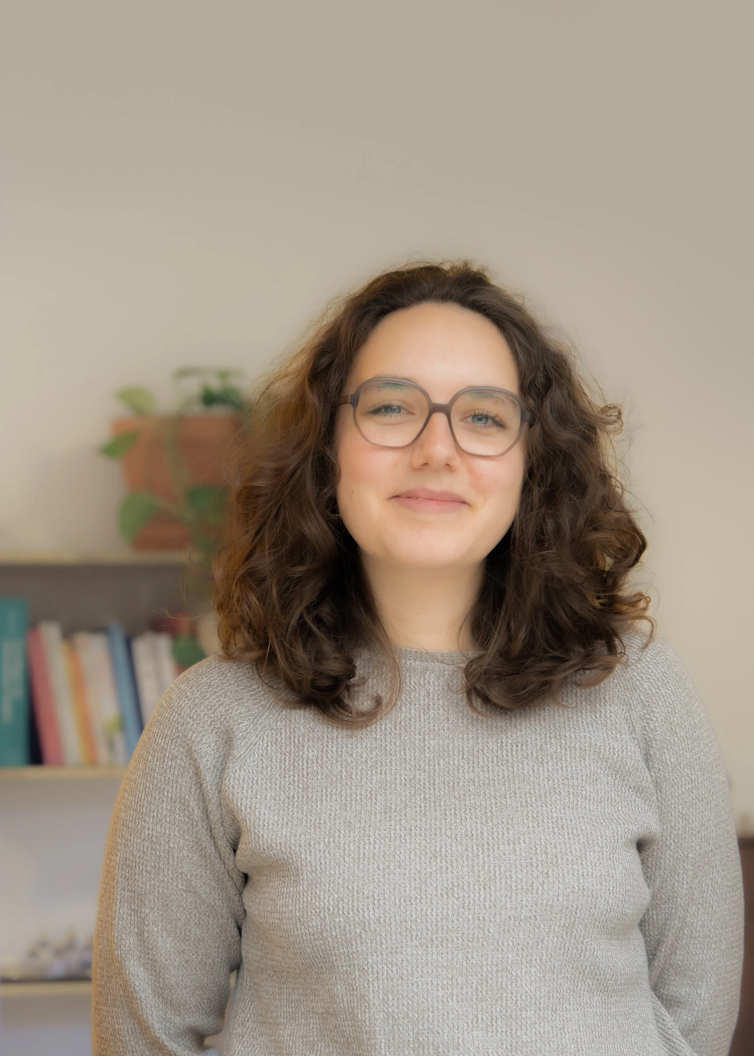 Porträt einer jungen Frau mit Brille, lockigem Haar und einem grauen Pullover, lachend, im Hintergrund ein Bücherregal und eine Pflanze.