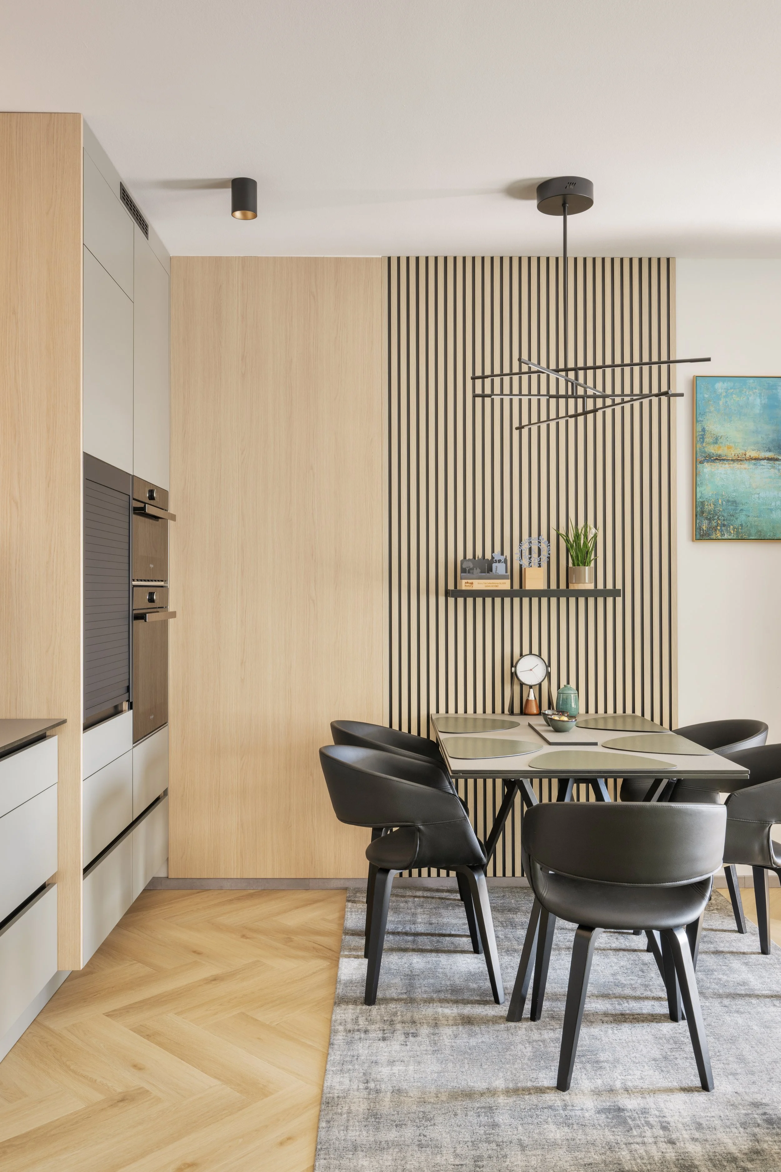 Interior of a modern dining room with a wooden wall, black dining table, and chairs, a shelf with decorative items and plants, artwork on the wall, and ceiling lights.