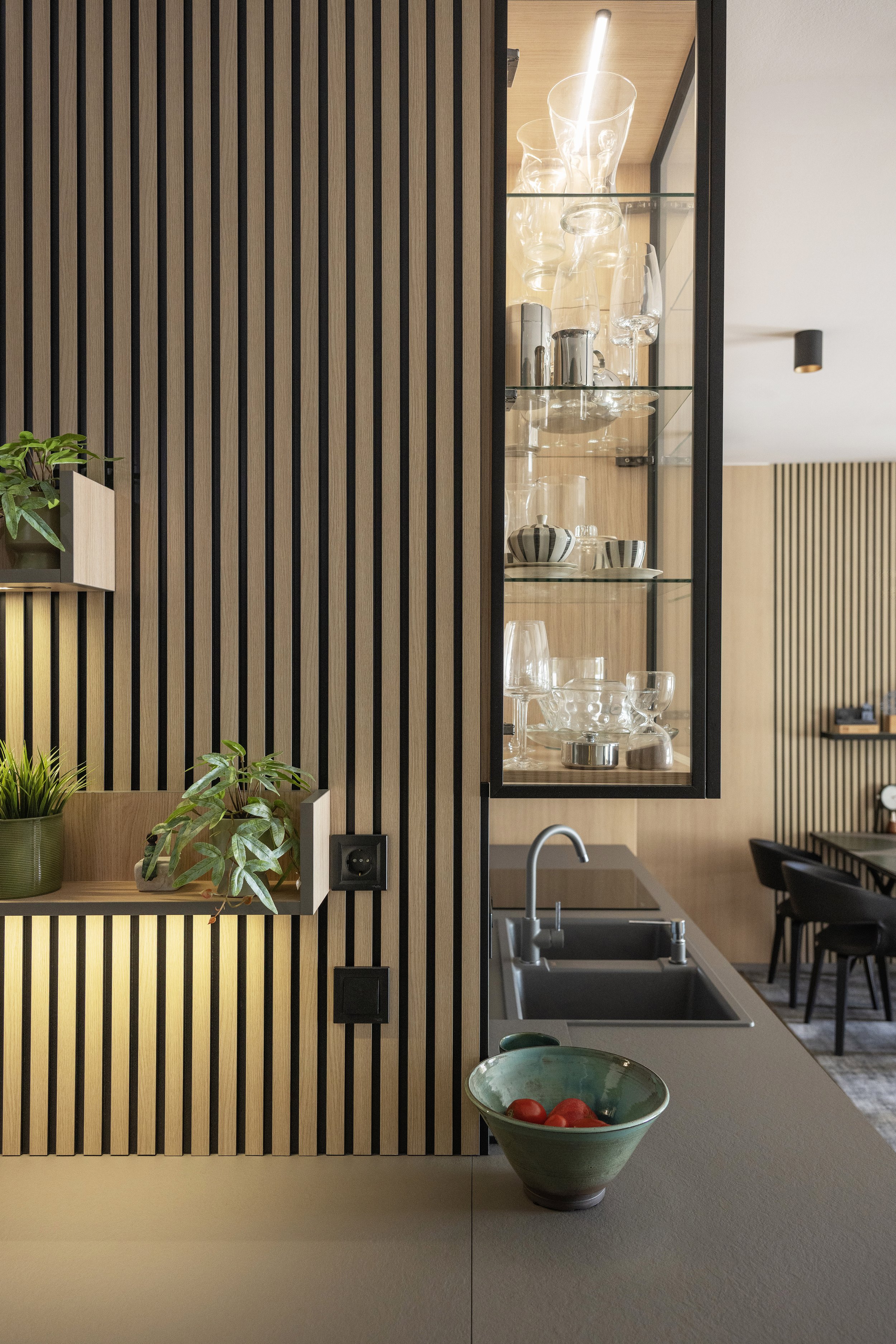 Modern kitchen interior with wooden slatted wall, open glass display cabinet with various glassware and ceramics, potted plants, bowl with red tomatoes on the countertop, black chairs, and a sink with a faucet