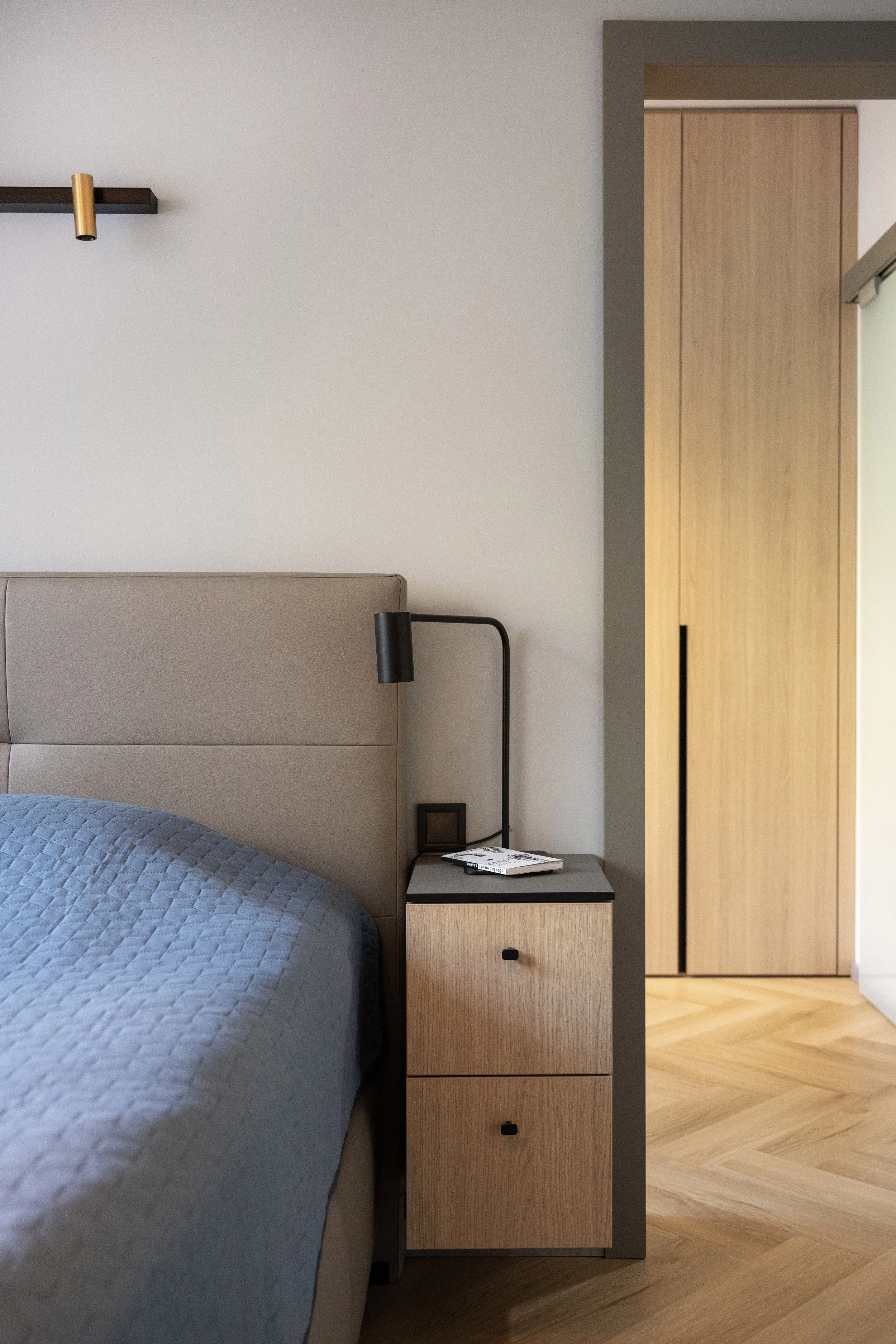 Bedroom with a bed covered in a blue quilt, a wooden nightstand with books and a reading light, a partial view of a closet, and light wood flooring.