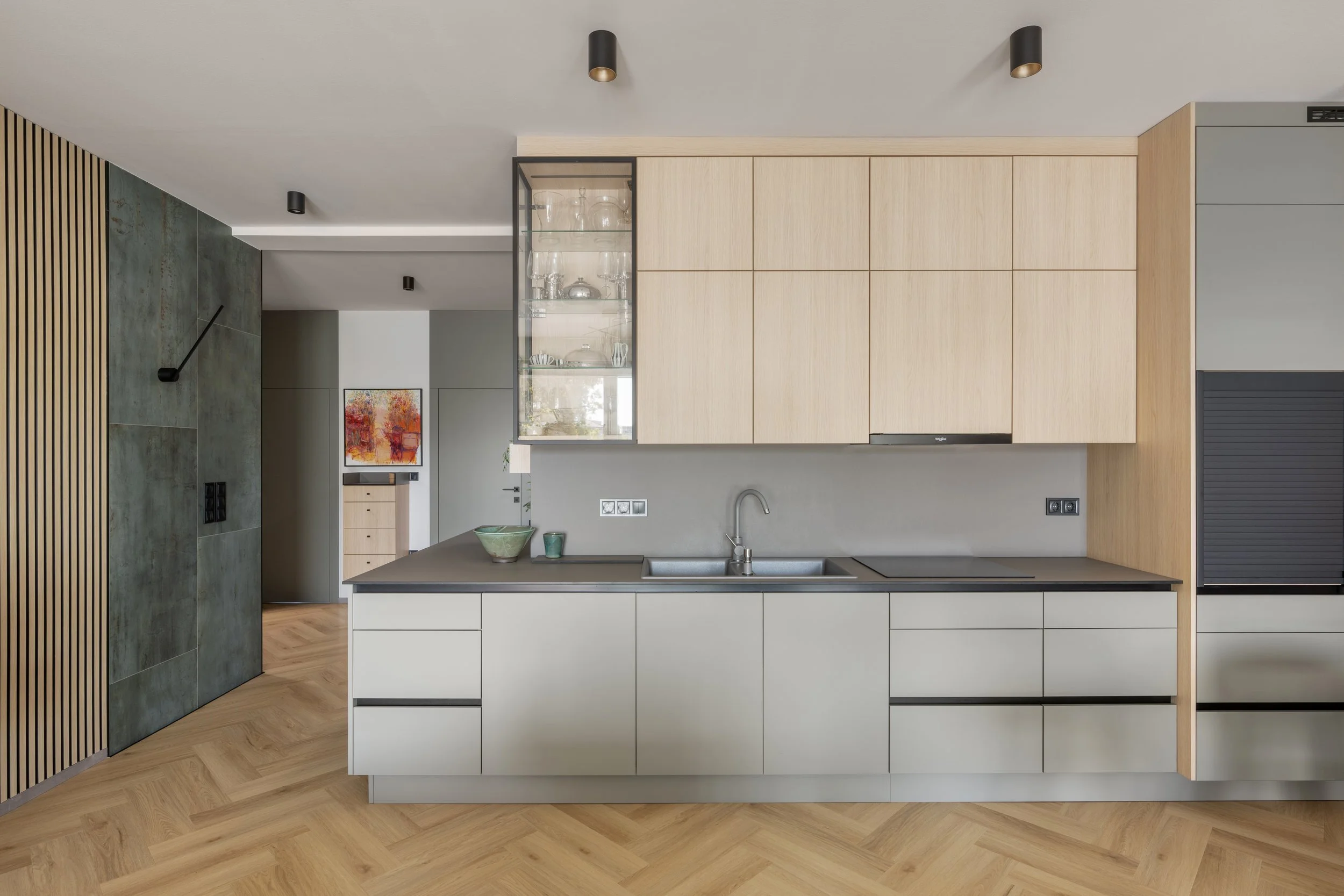 Modern kitchen with light wood cabinets, black countertops, gray and wood-paneled wall, and a glass display cabinet with glassware, against a backdrop of white walls and warm wooden flooring.