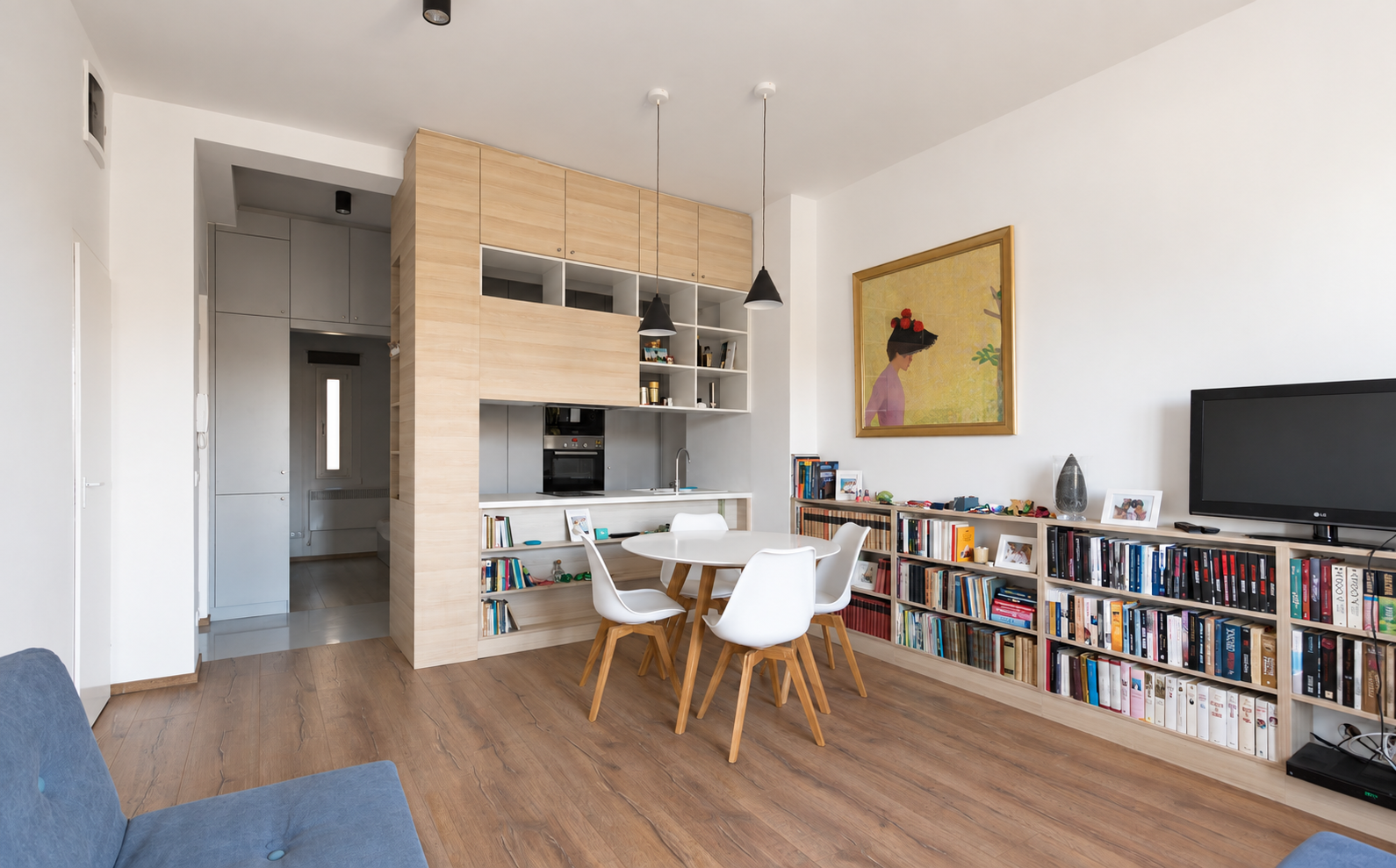 Modern living room with wooden flooring, white walls, a round dining table with four white chairs, a bookshelf filled with books, a flat-screen TV, and framed artwork on the wall.