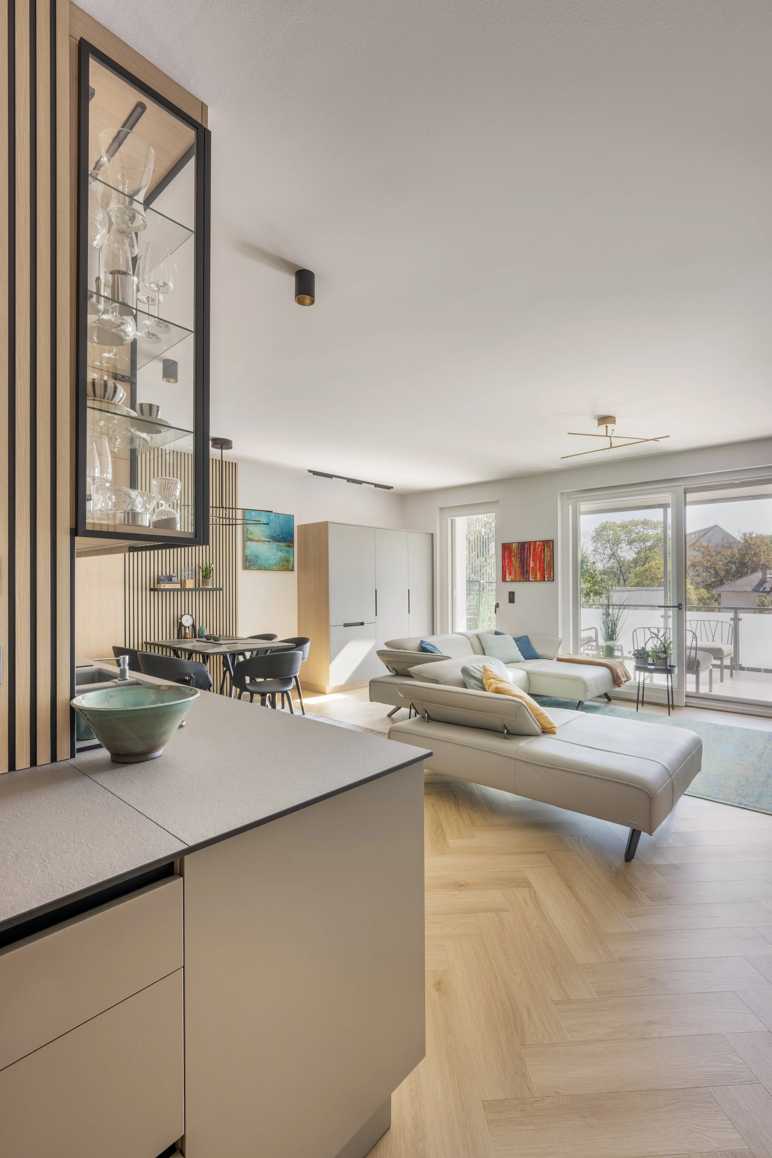 Modern living room with white sofa, colorful throw pillows, wooden floors, large windows, and a dining area with black chairs and a light-colored table. Glass cabinet with glassware decorates the corner.