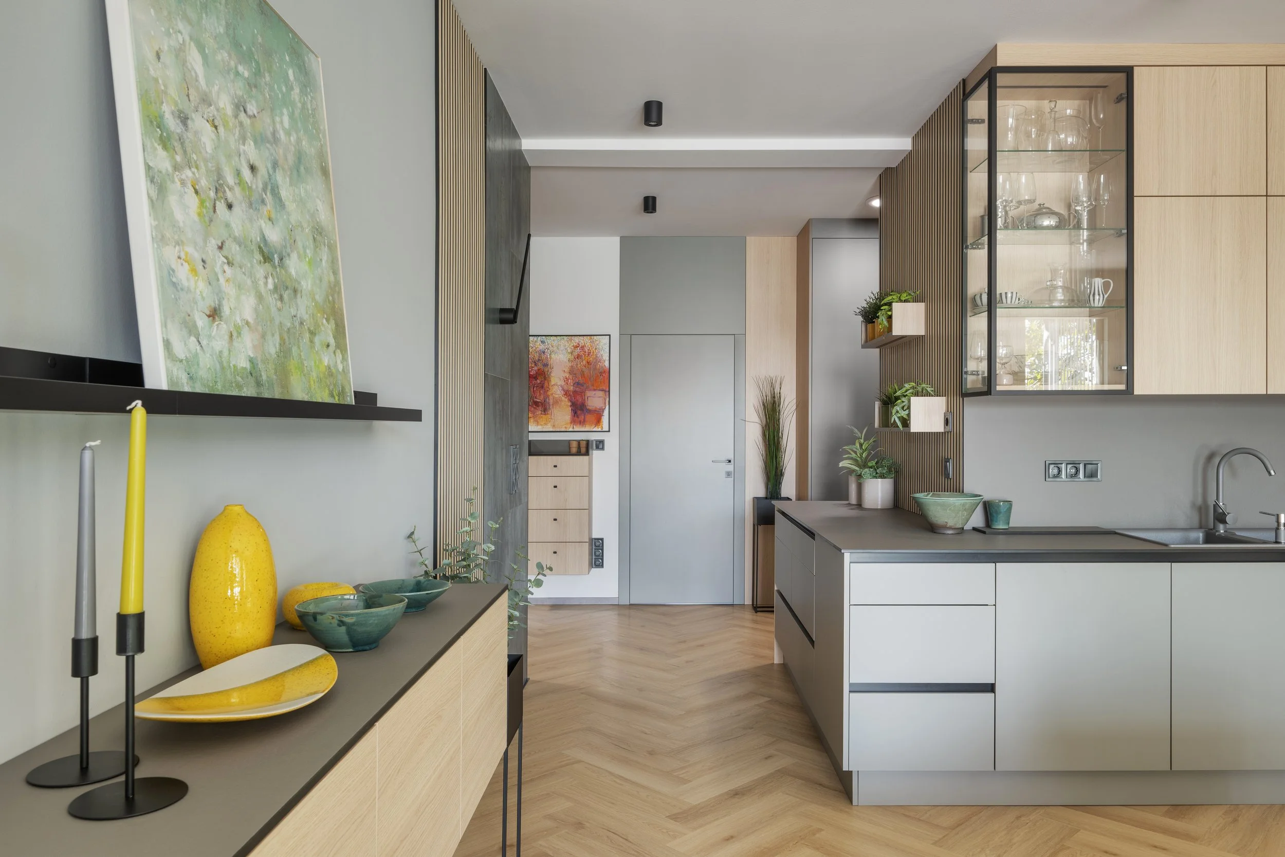 Modern kitchen with light wood cabinets, gray countertops, and decorative vases and bowls, with abstract paintings on the wall and a wooden slat wall feature.