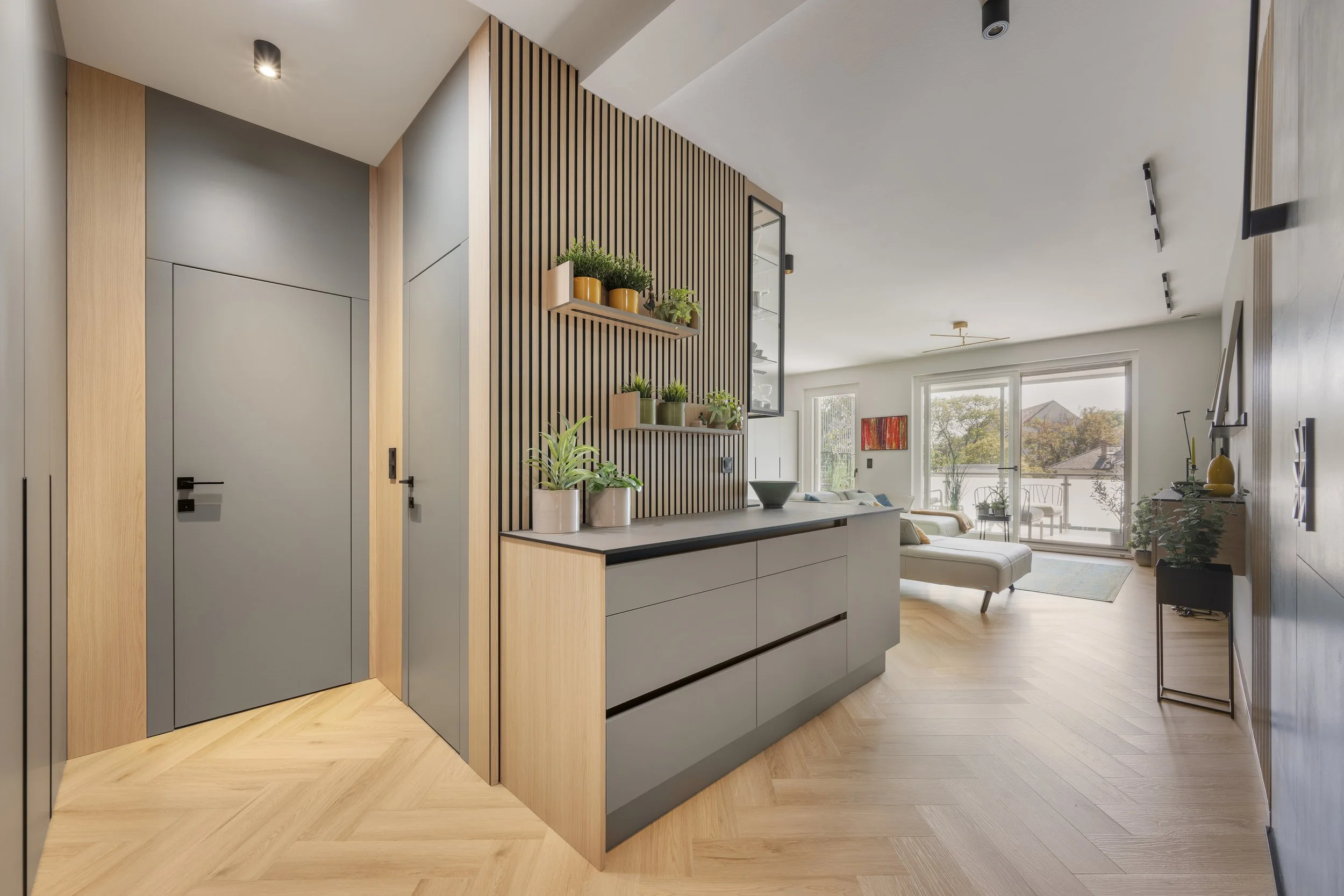 Modern interior living space with a partial view of a kitchen and a lounge area, featuring wood flooring, a slatted wood accent wall with floating shelves holding potted plants, and large windows or sliding doors letting in natural light.